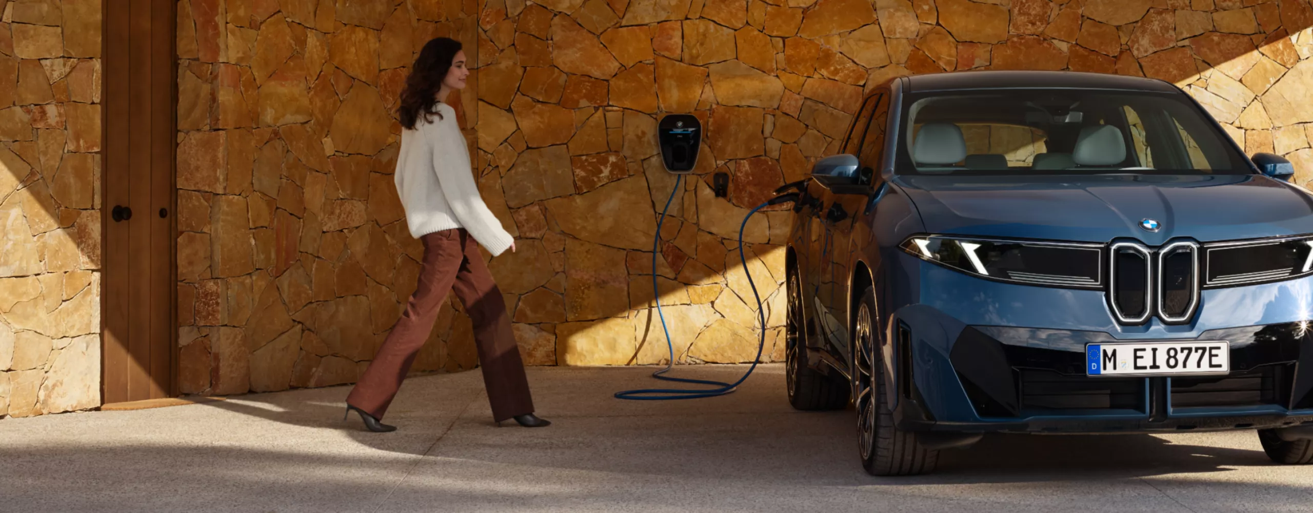 A woman approaches a fully electric BMW, which is connected to a charging point on a house