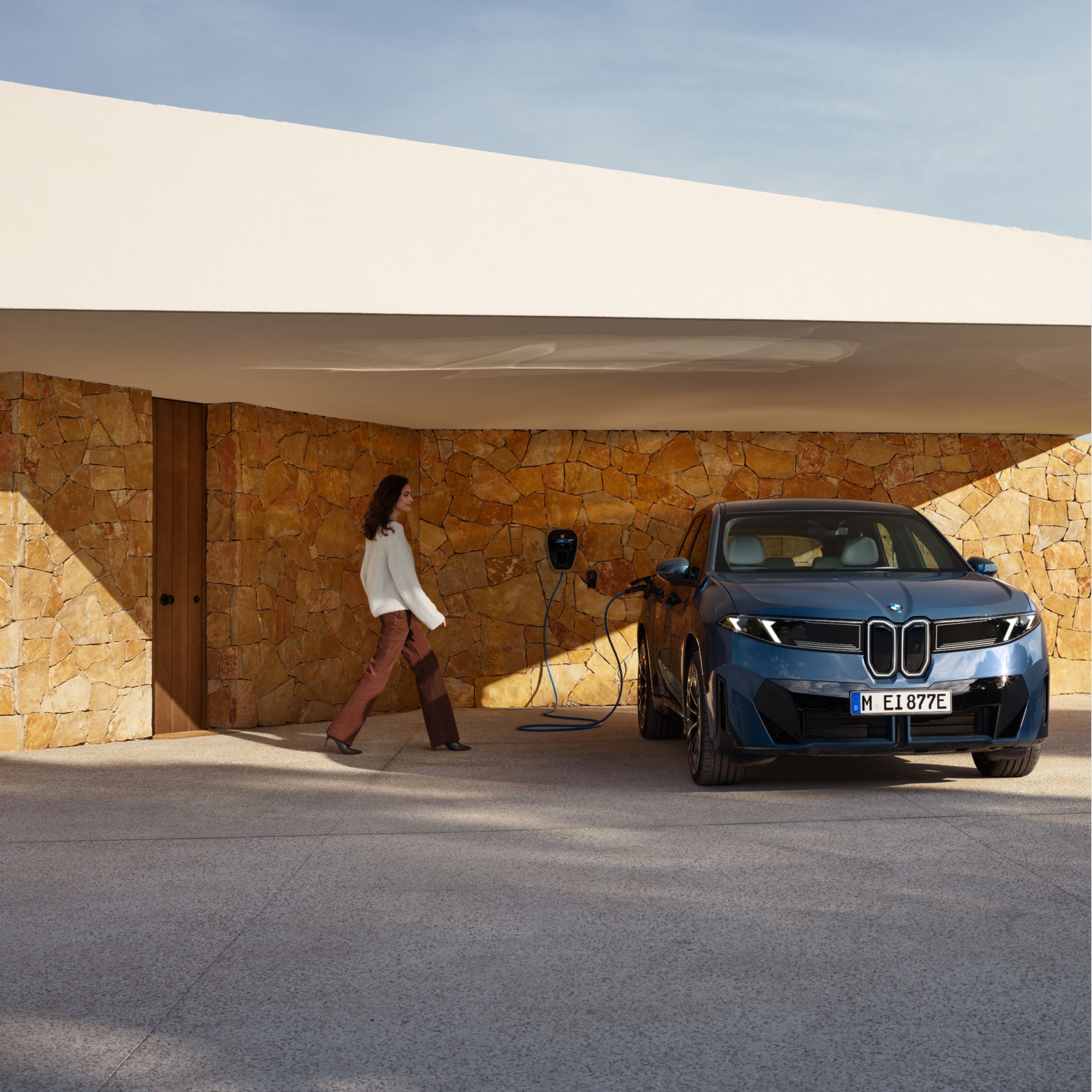 A woman approaches a fully electric BMW, which is connected to a charging point on a house