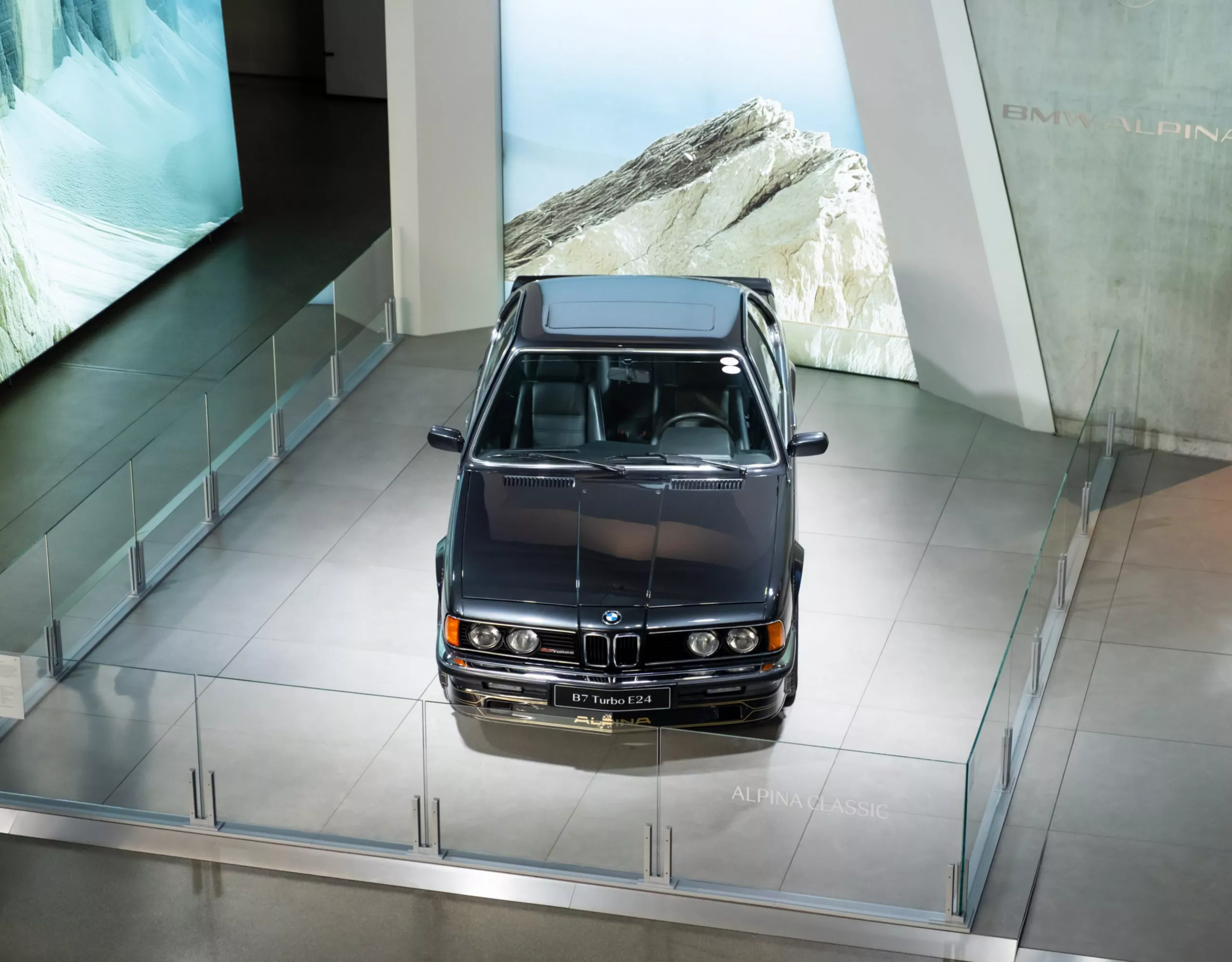 Alpina B7 Turbo (E24) displayed at BMW Welt on a glass-enclosed platform in a modern exhibition space with illuminated mountain landscape panels.