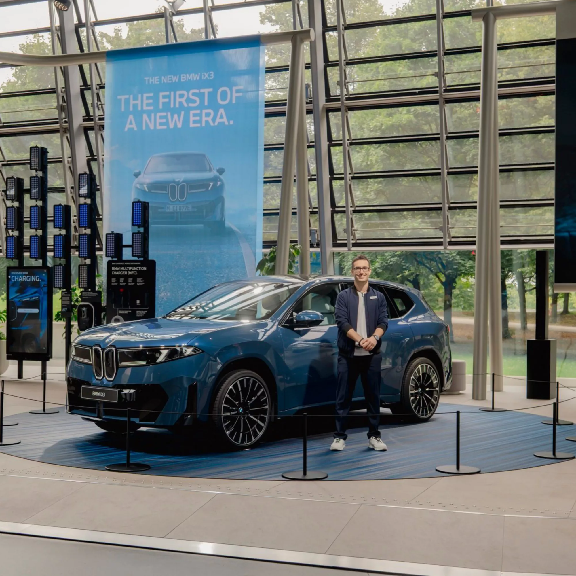 BMW 7 Series Sedan in a two-tone paint finish of Oxid Grey and Sapphire Black, on display at BMW Welt against a backdrop of modern glass architecture.