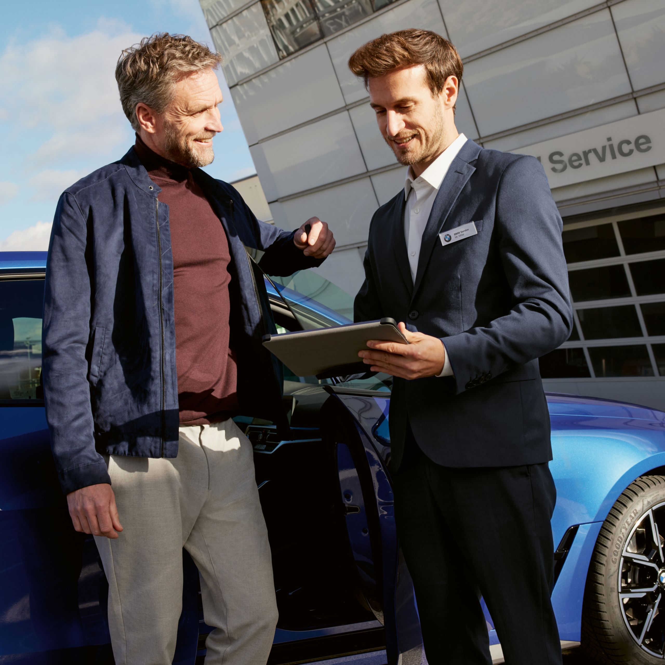 Kunde steht vor seiner geöffneten Autotür (blauer BMW) neben einem BMW Mitarbeiter, der ein Tablet in der Hand hält. Im Hintergrund ist ein Gebäude mit dem Schild BMW Service zu erkennen.