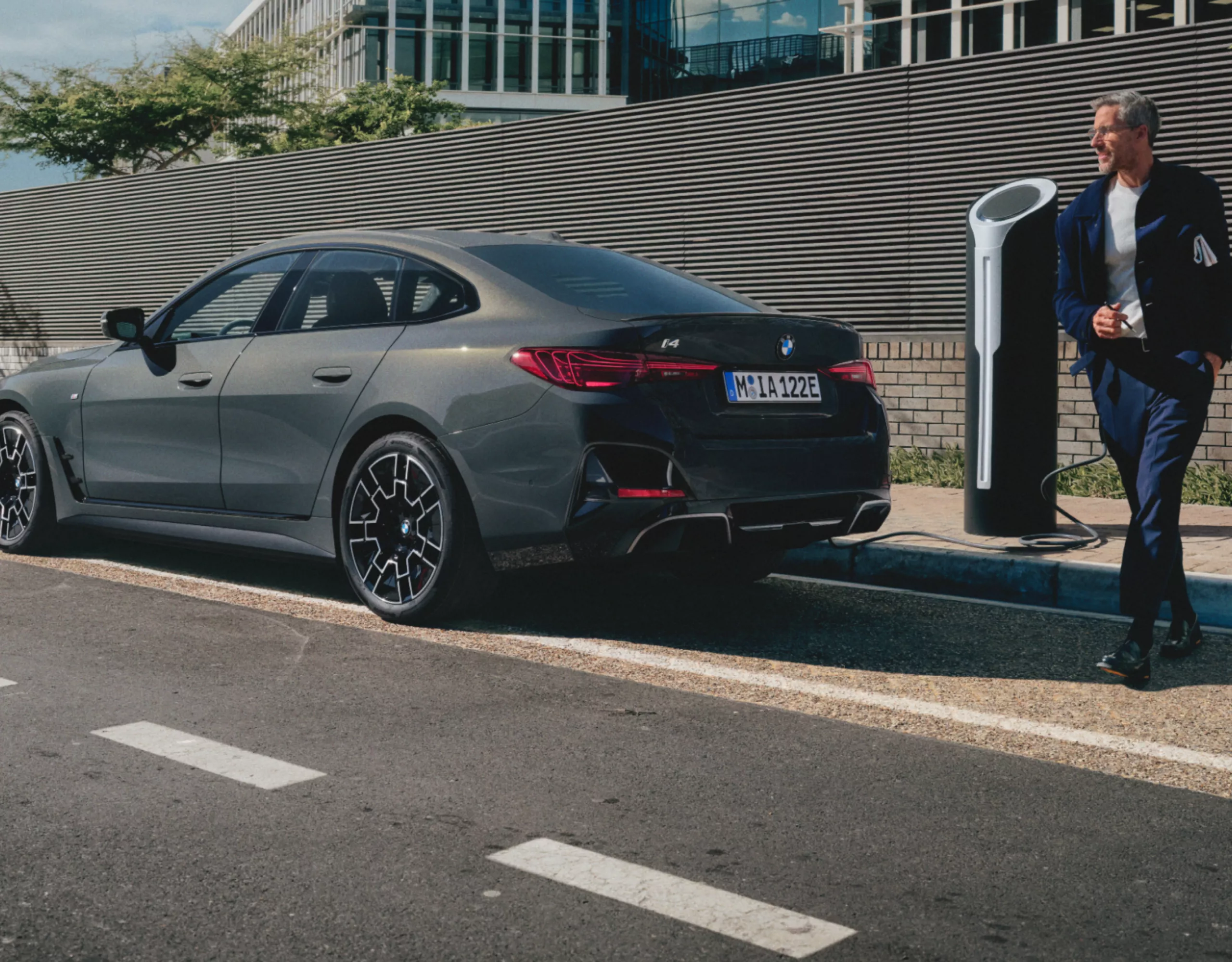 Ein grauer BMW und ein Mann stehen an der Straße vor einer Mauer. Der BMW lädt an einer Ladesäule