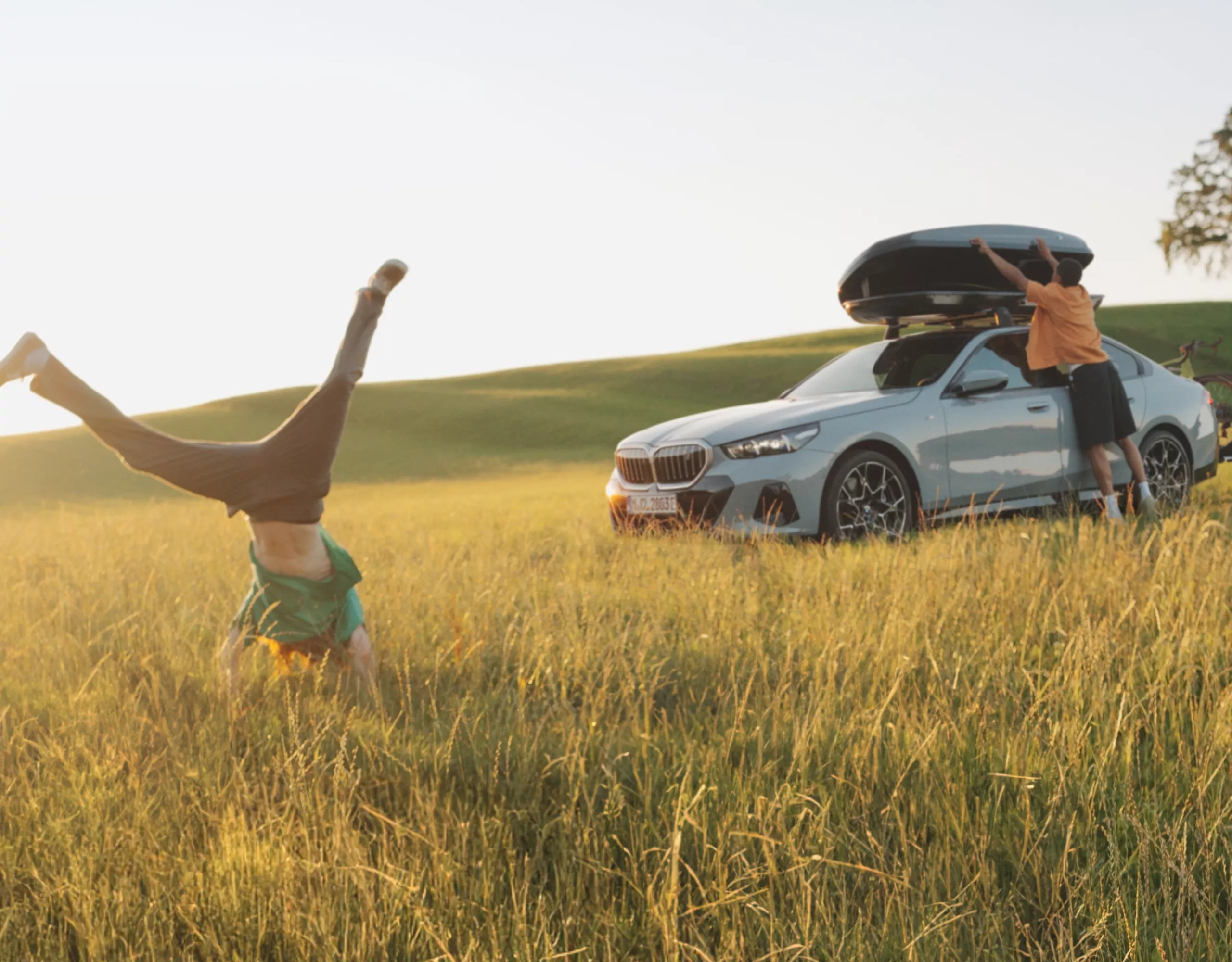 A person does a handstand on a meadow in summer. A BMW with a roof box is parked in the background