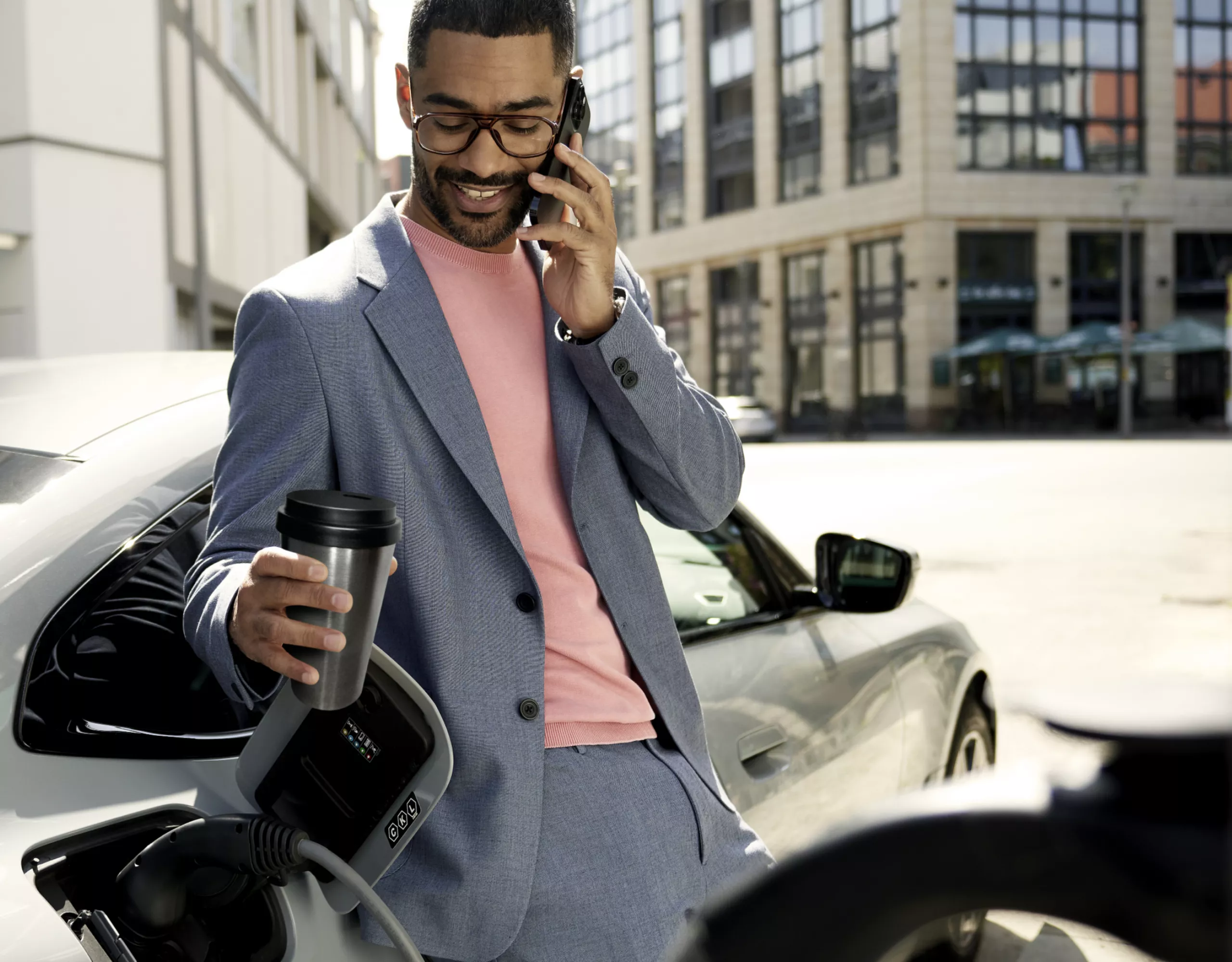 Lächelnder Mann telefoniert vor einem vollelektrischen BMW an einer Ladesäule und trinkt Kaffee 