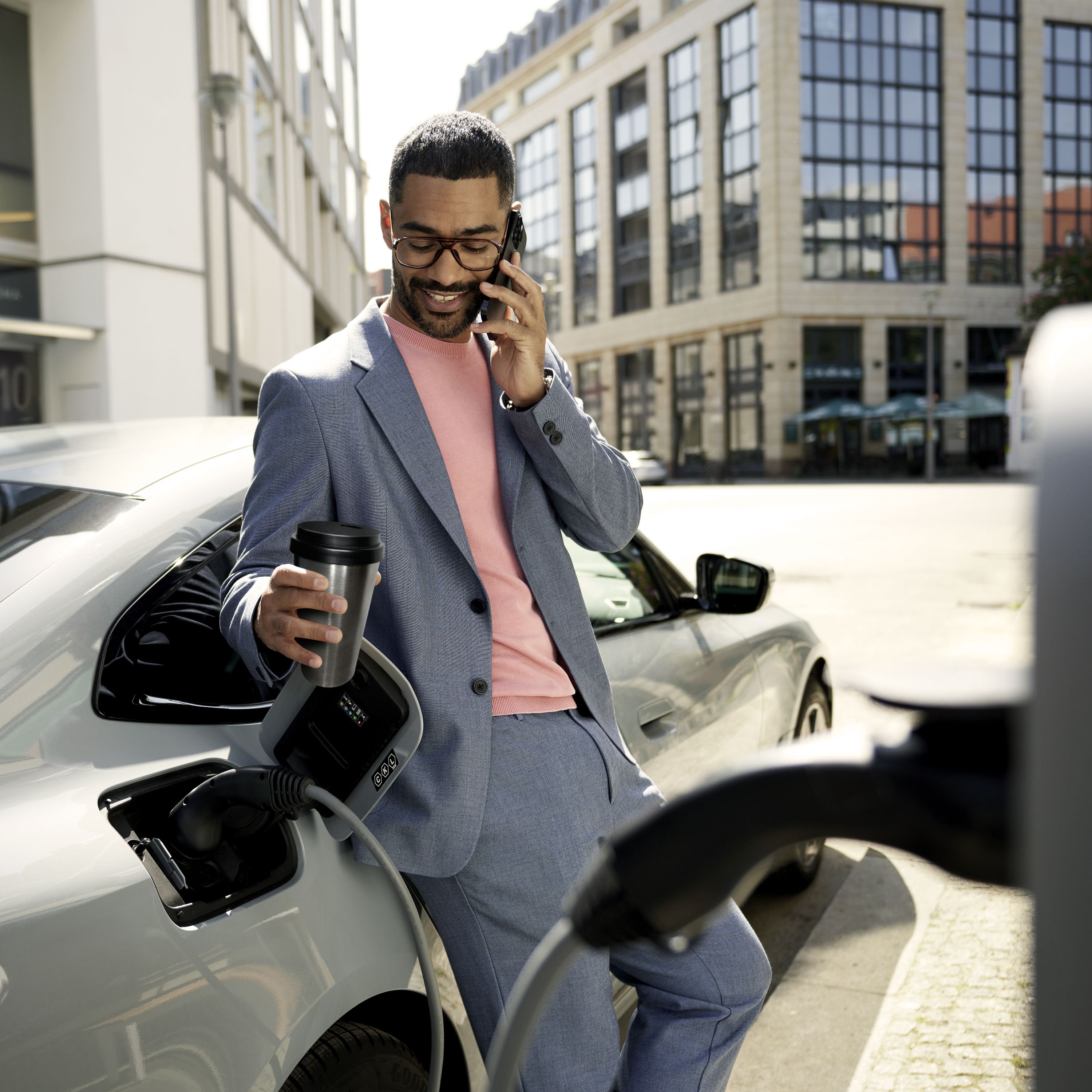 A smiling man is having a phone call in front of a BMW connected to a quick charger while drinking coffee