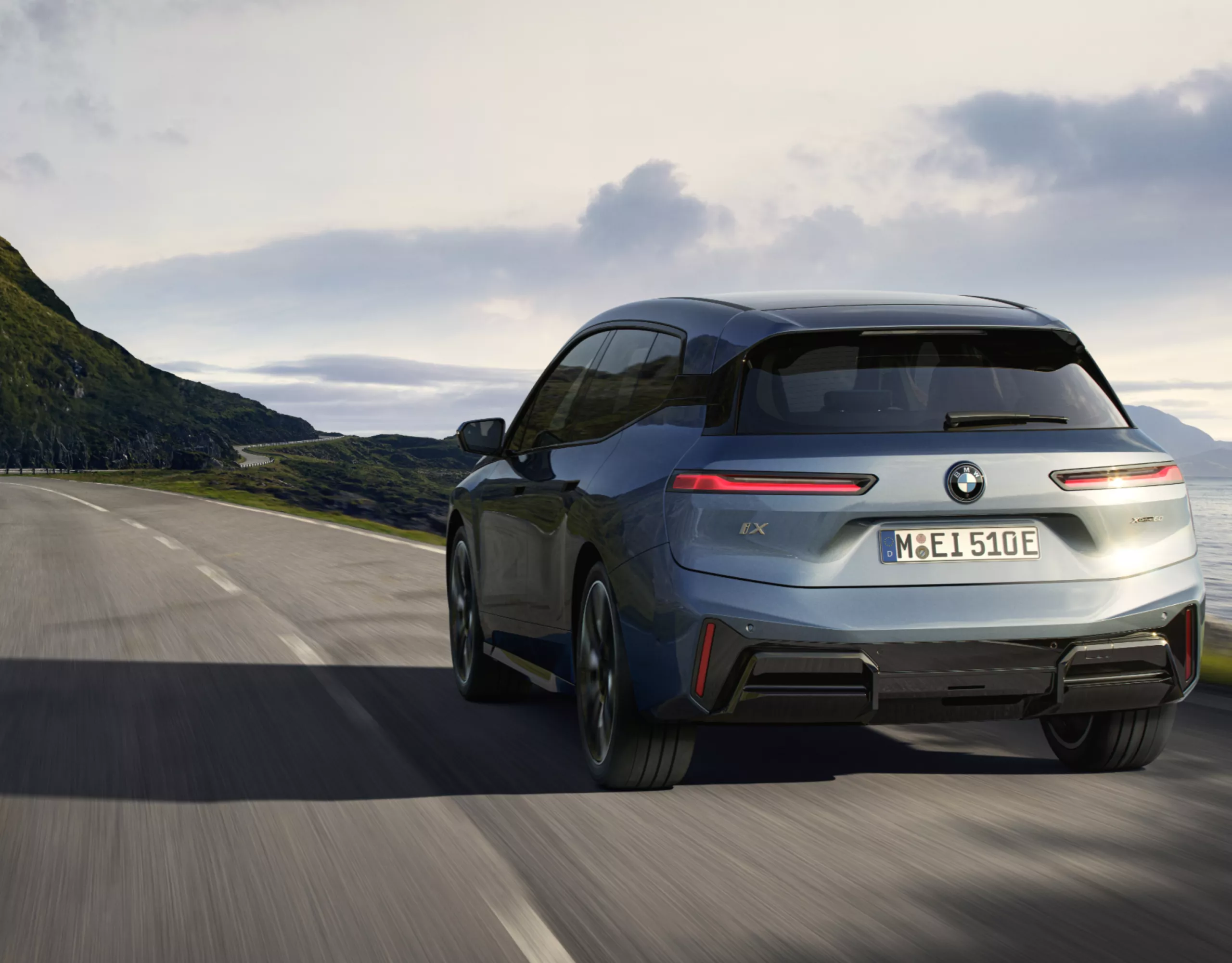 Ein BMW iX in hellblau fährt auf einer Straße am Meer in Richtung Berge