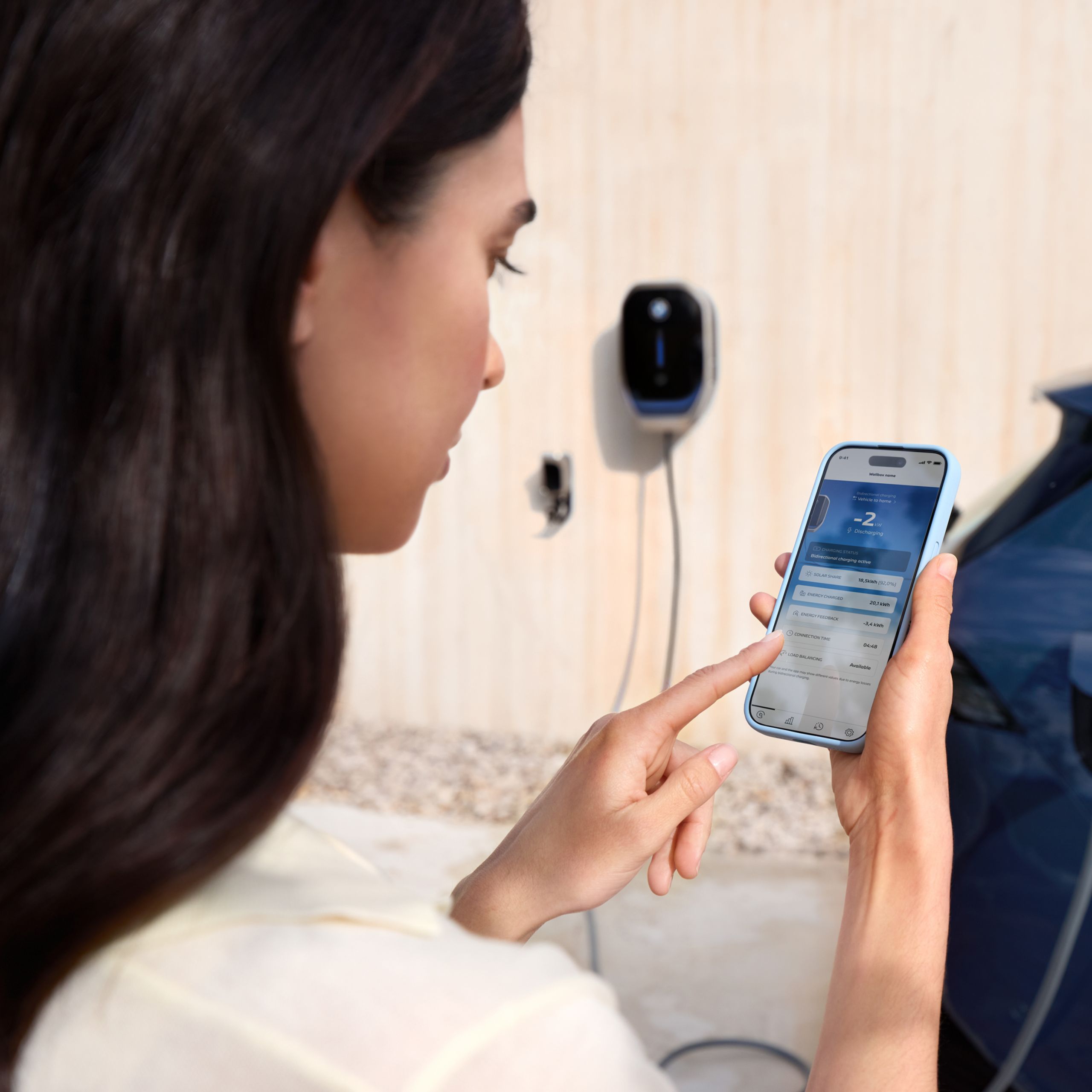 Eine Frau blickt auf Ihr Smartphone, im Hintergrund ist eine BMW Wallbox