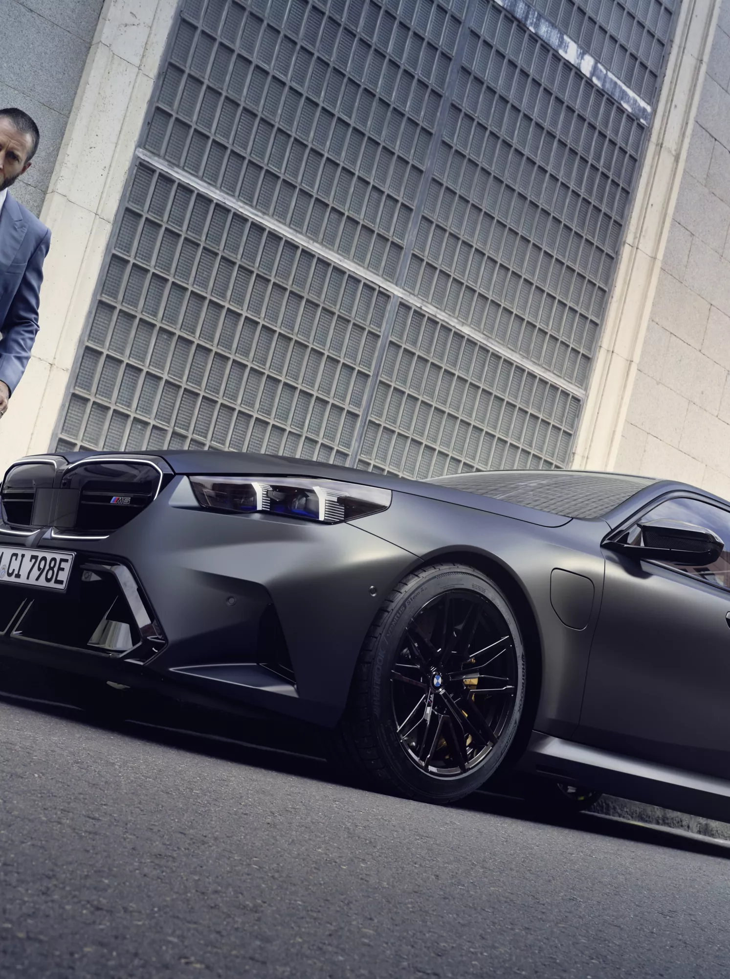 Tyylikkäästi pukeutunut mies ohittaa pysäköidyn harmaan BMW M5-mallin.