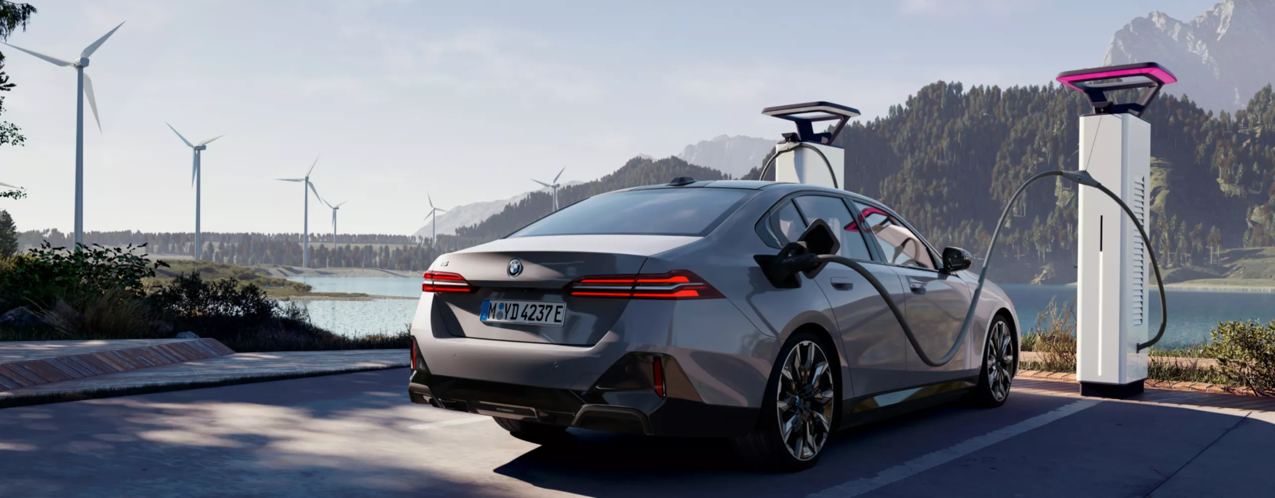 BMW laadt op bij een laadstation met uitzicht op water, bergen en windturbines