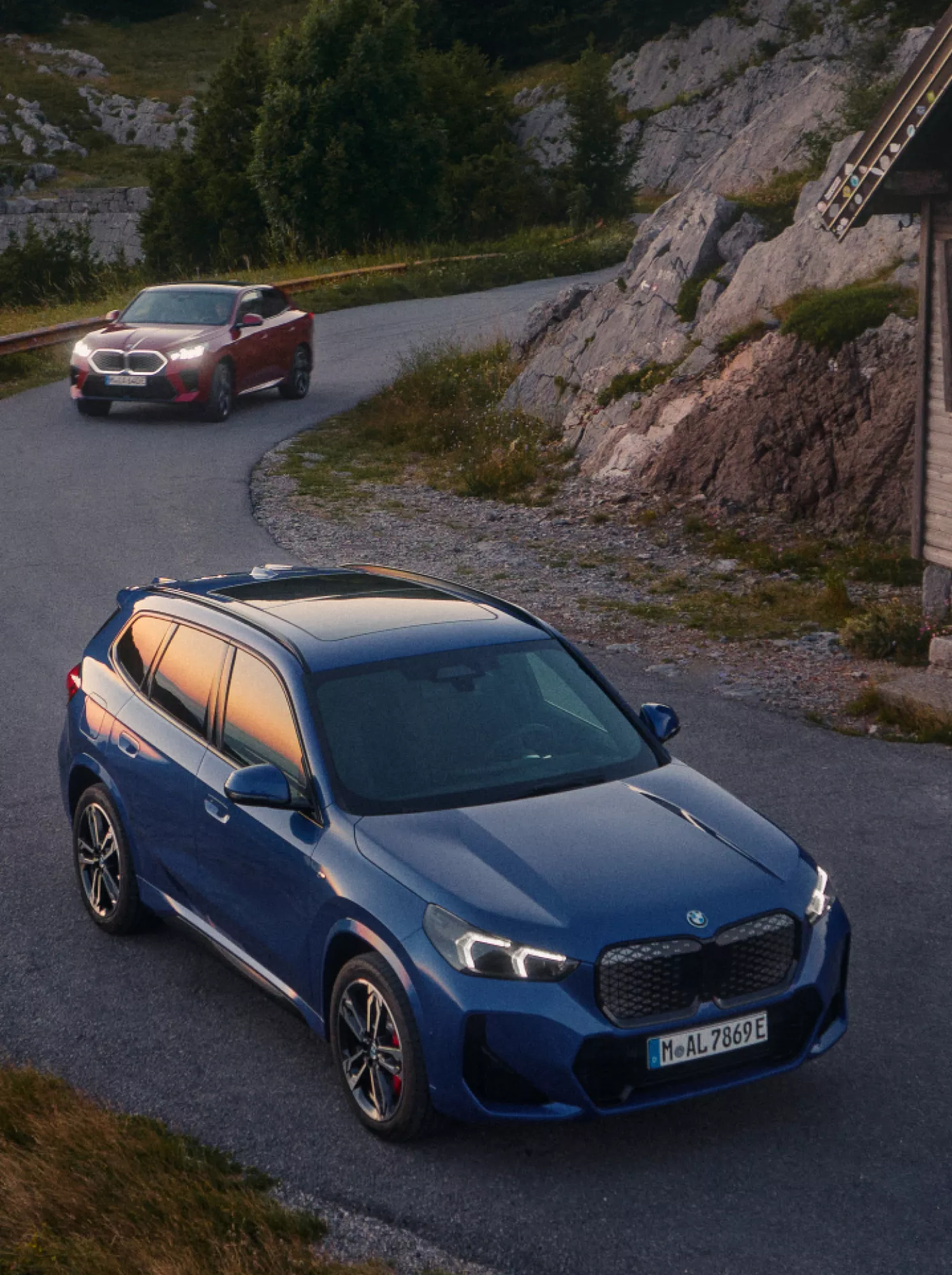 A BMW iX1 and iX2 on a mountain road in front of a wooden hut