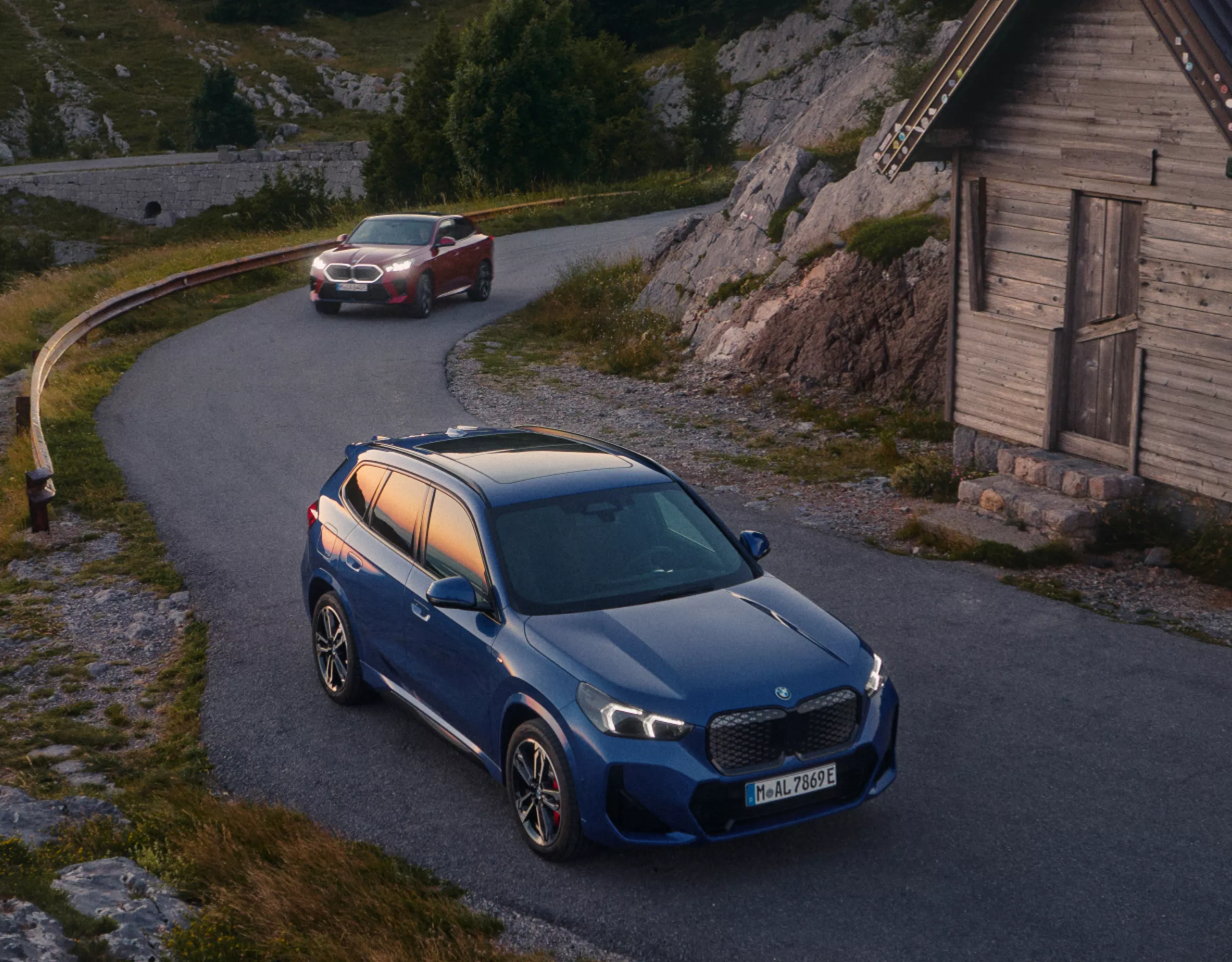木造小屋の前の山道を走るBMW iX1とBMW iX2