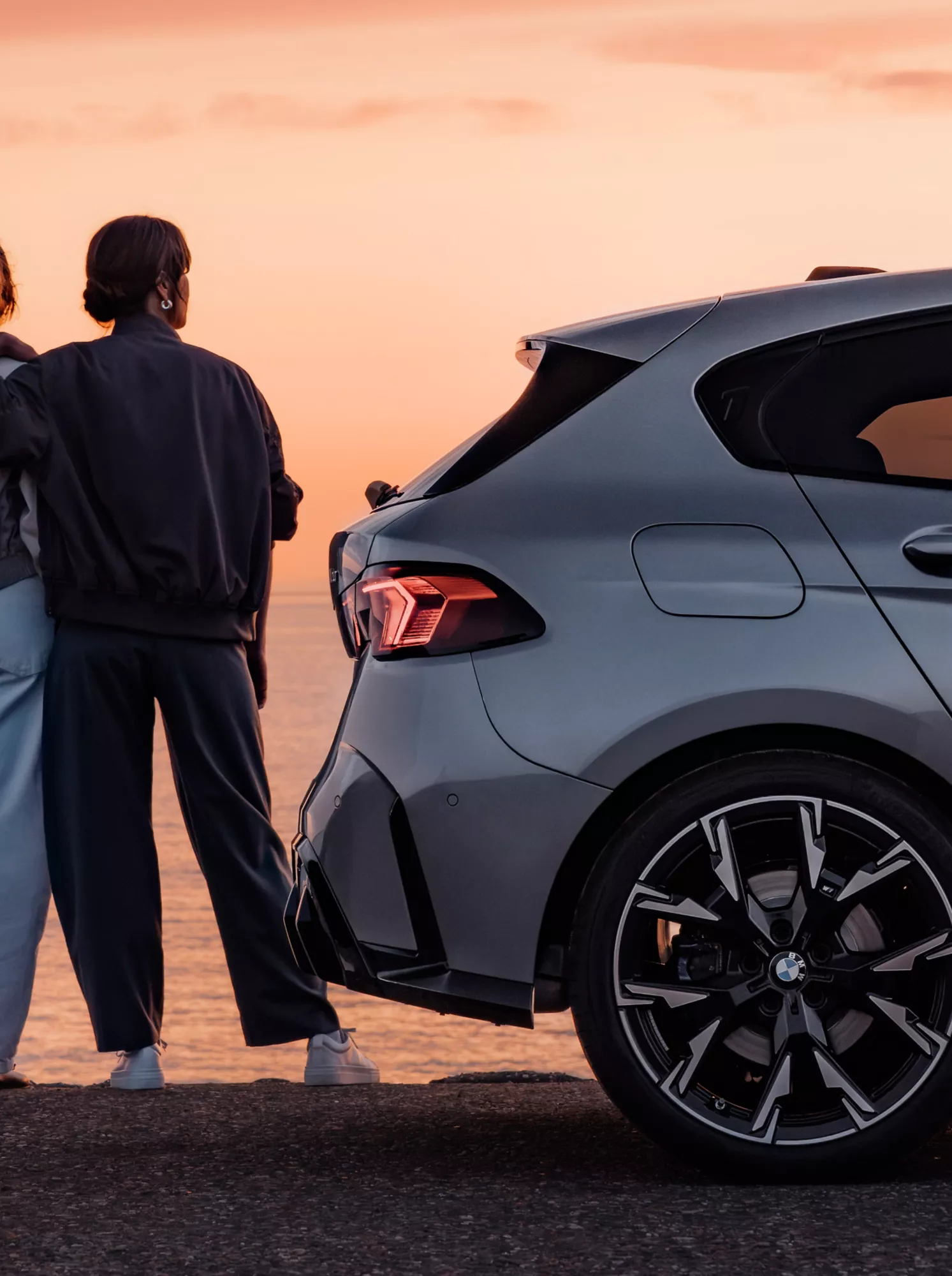 Vista posteriore di una BMW Serie 1 parcheggiata sulla costa al tramonto, con due persone accanto che ammirano il panorama