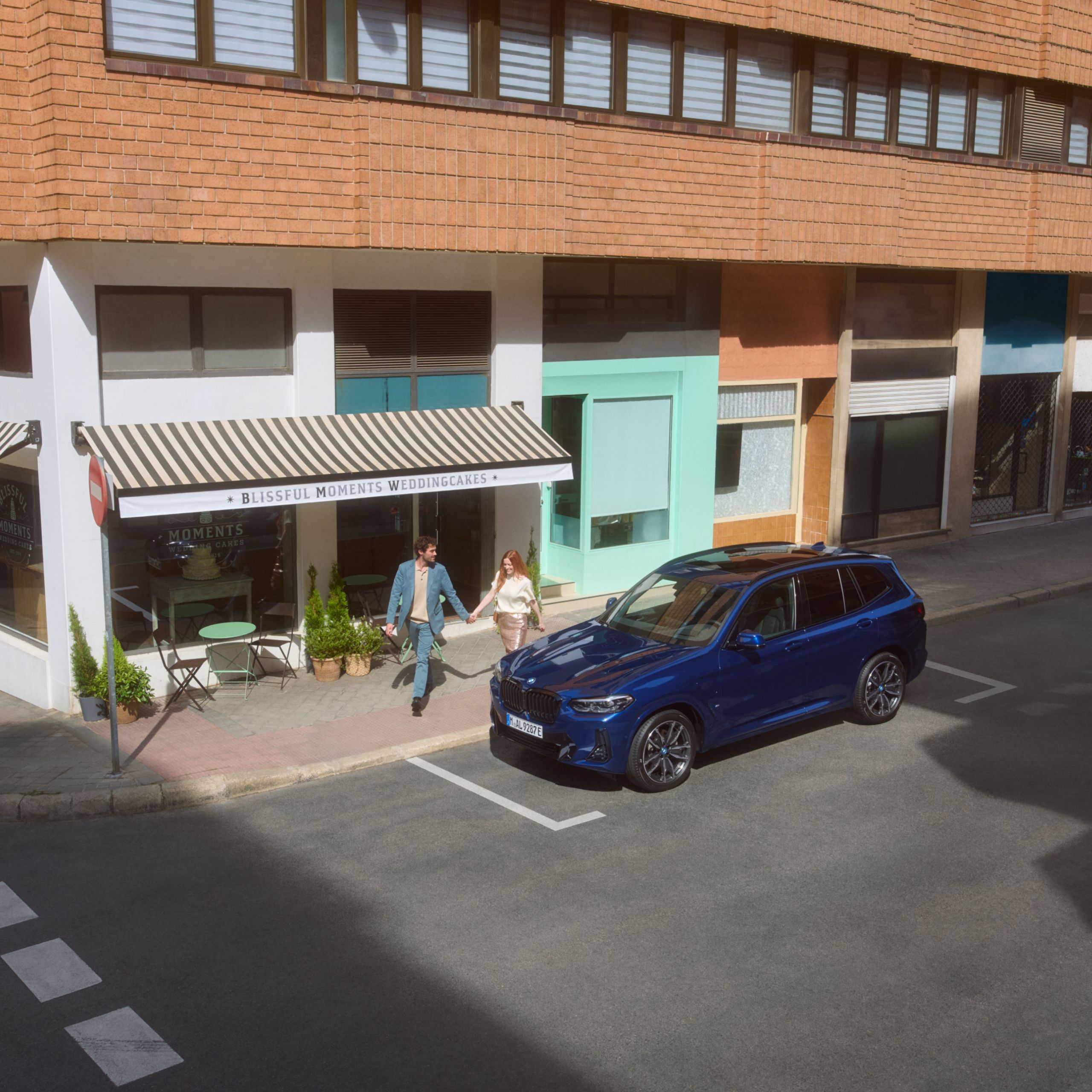 Dvaja ľudia kráčajú po chodníku a držia sa za ruky. Pri ceste je zaparkované BMW