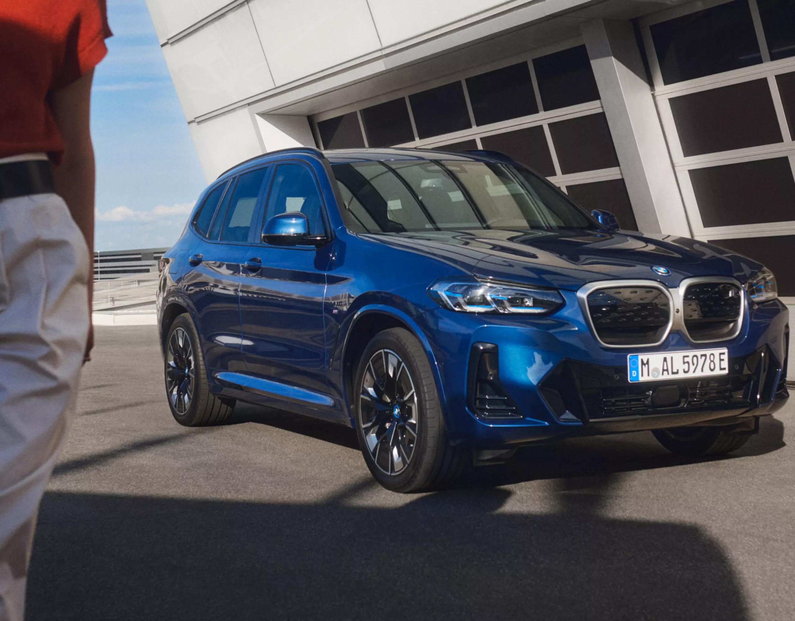 Vor einem Gebäude steht ein BMW. Eine Person läuft teilweise aus dem Bild
