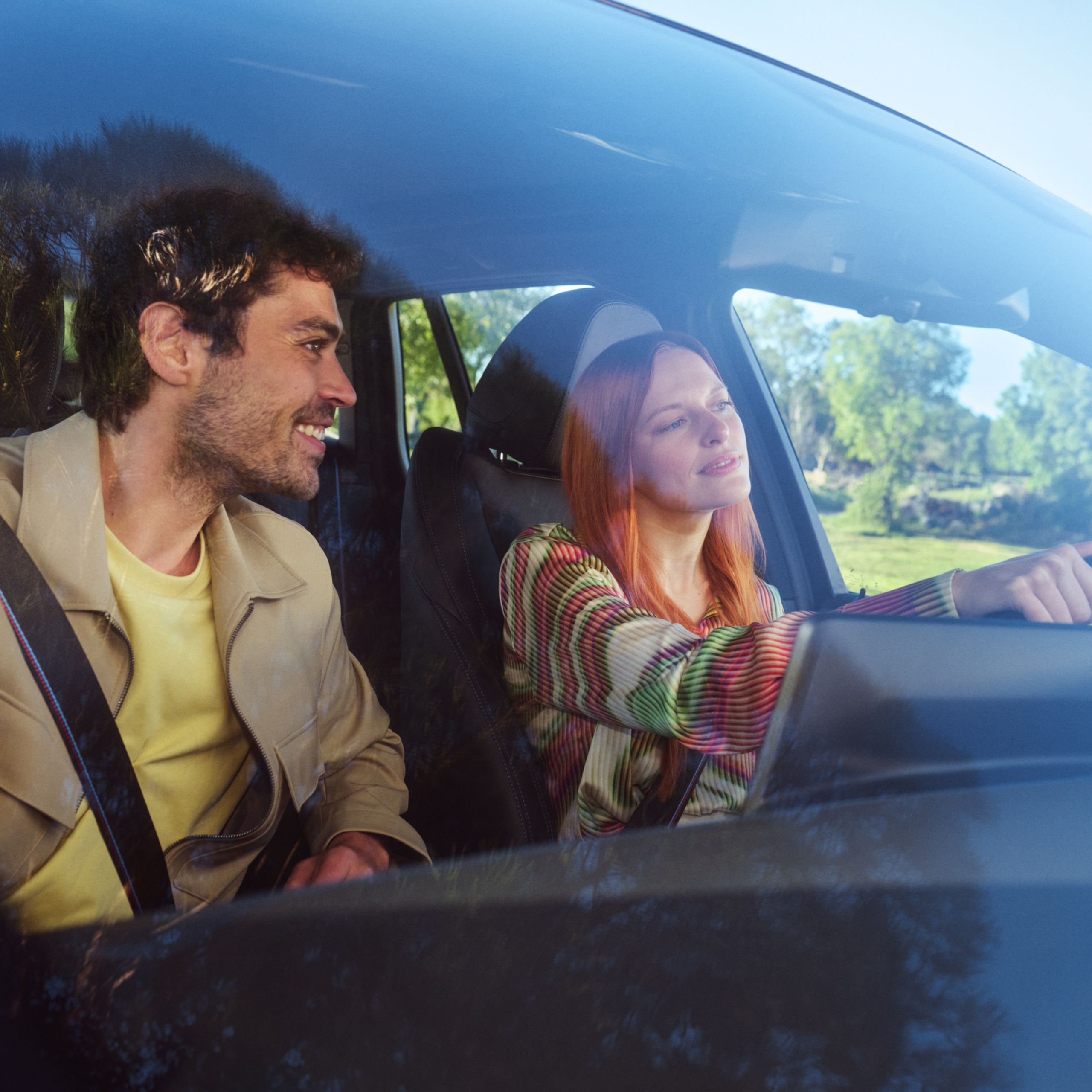 Cez čelné sklo vidieť ženu, ktorá šoféruje, zatiaľ čo muž sa na ňu pozerá zo sedadla spolujazdca a usmieva sa