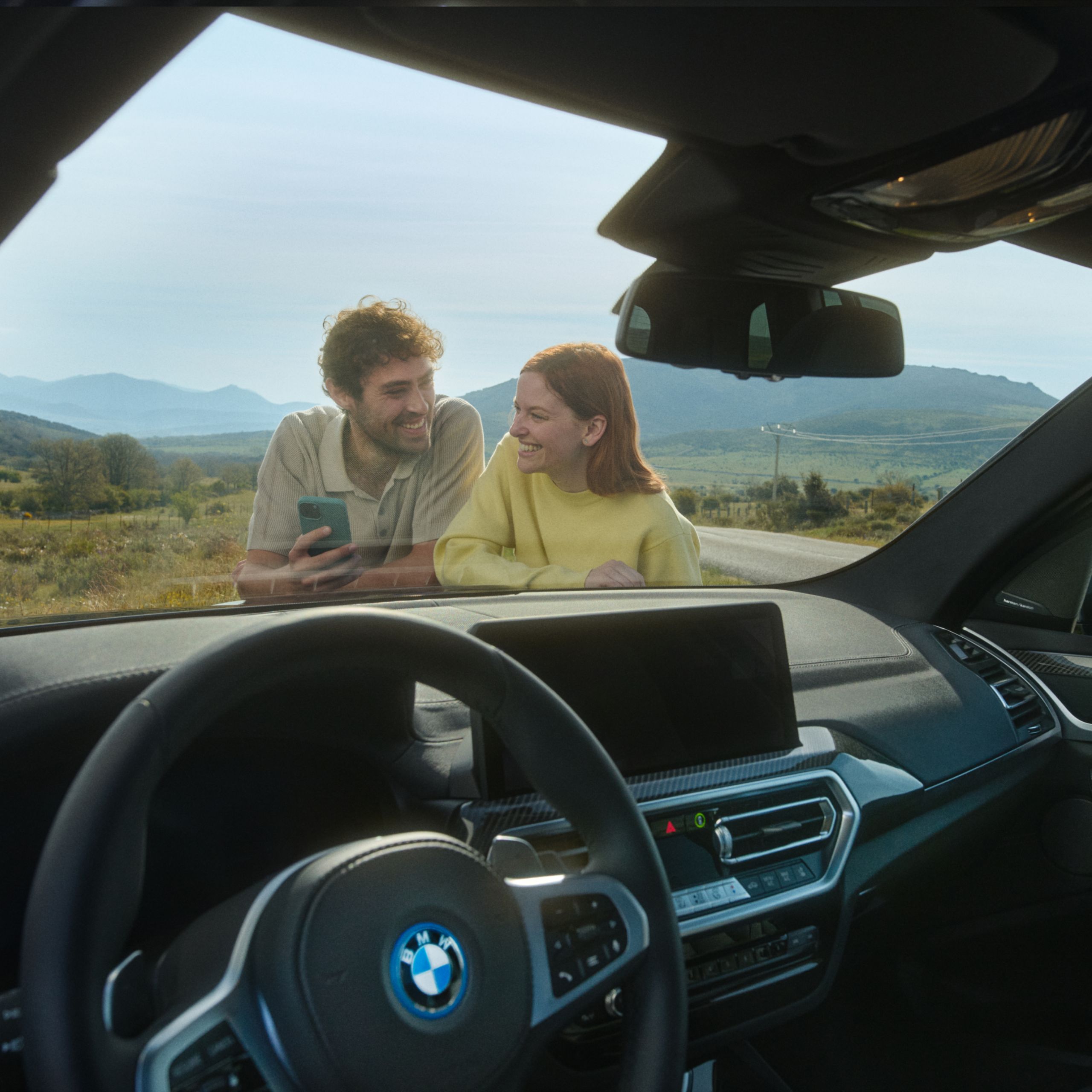 Cez čelné sklo vidieť ženu a muža, ktorí sa opierajú o kapotu a usmievajú sa. Muž drží smartfón