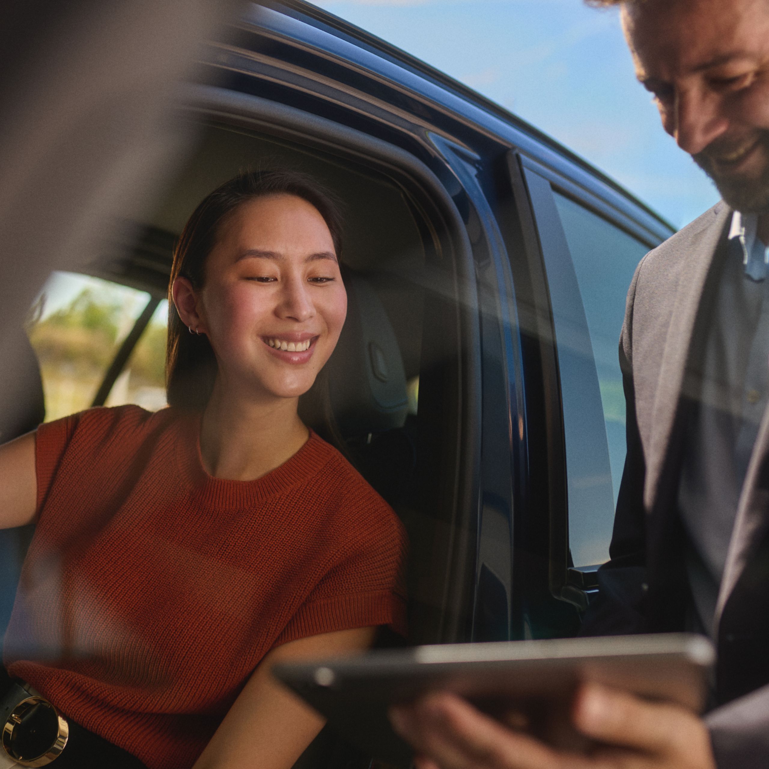 Žena sedí v BMW s otvorenými dverami. Vedľa nej je muž s tabletom. Obaja sa naň pozerajú a usmievajú sa