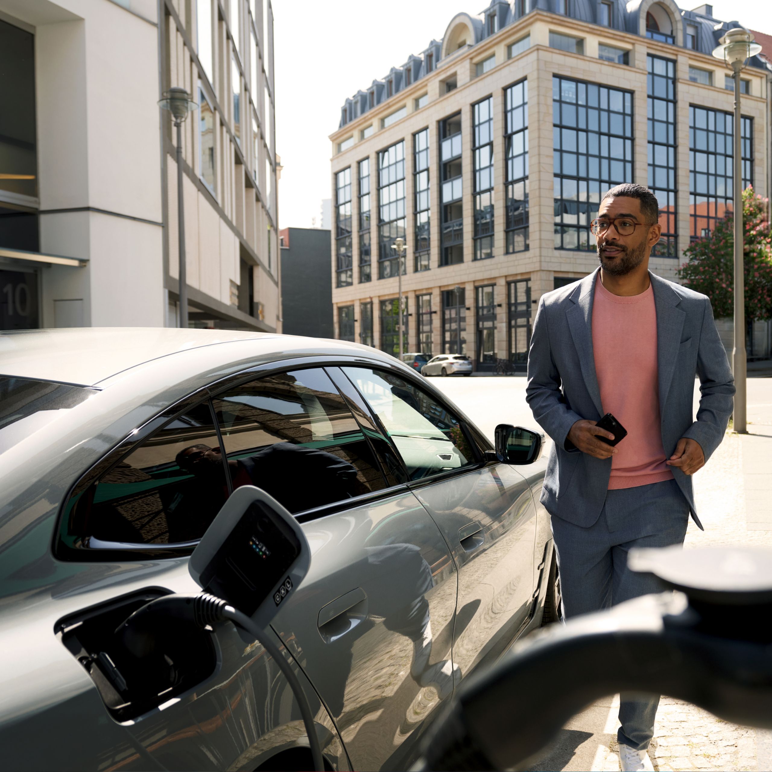 Muž držiaci smartfón prechádza okolo BMW, ktoré sa nabíja. V pozadí sú viditeľné budovy