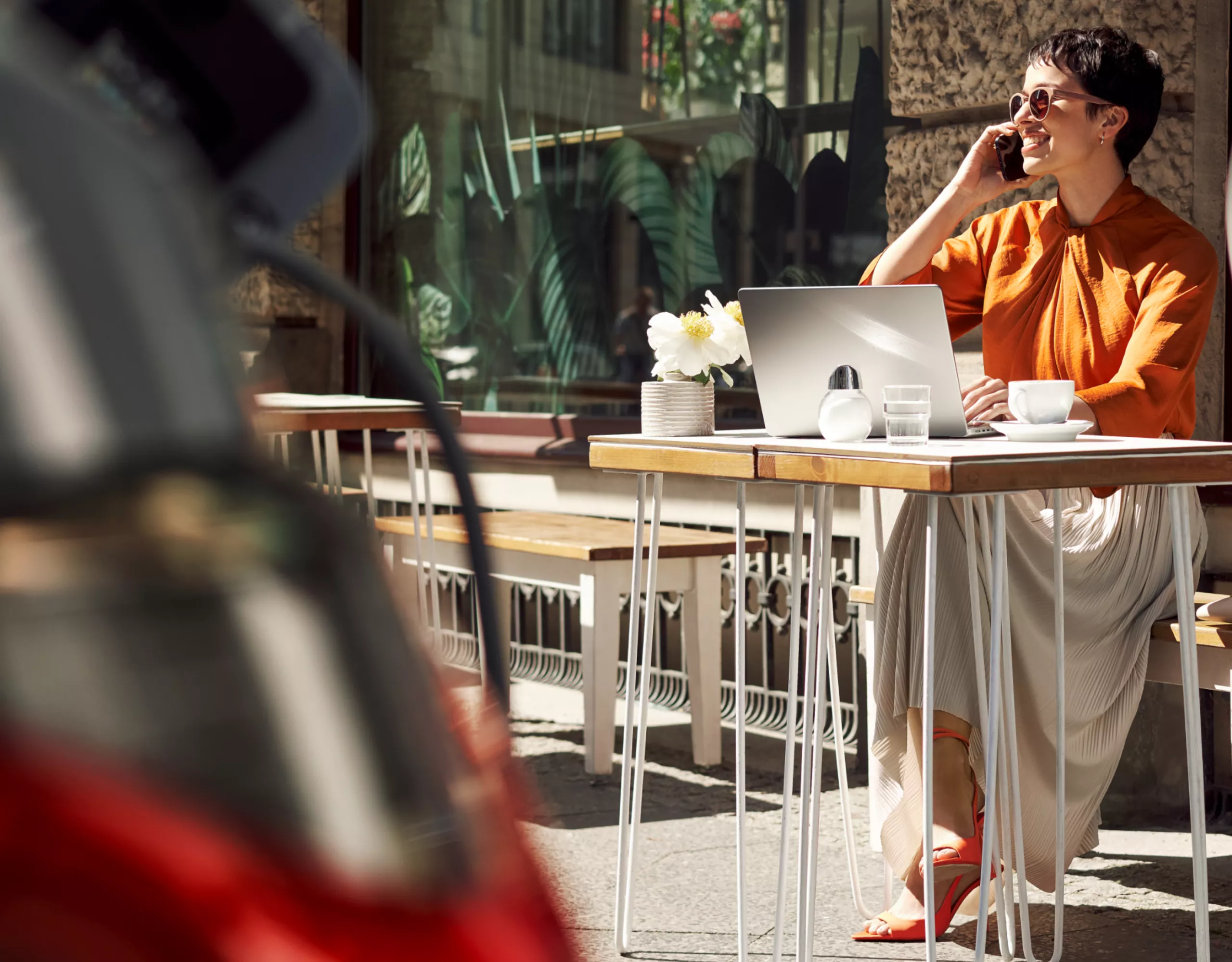 Vor einem Café sitzt eine Frau an einem Tisch mit offenem Laptop und telefoniert. Im Vordergrund lädt ein BMW