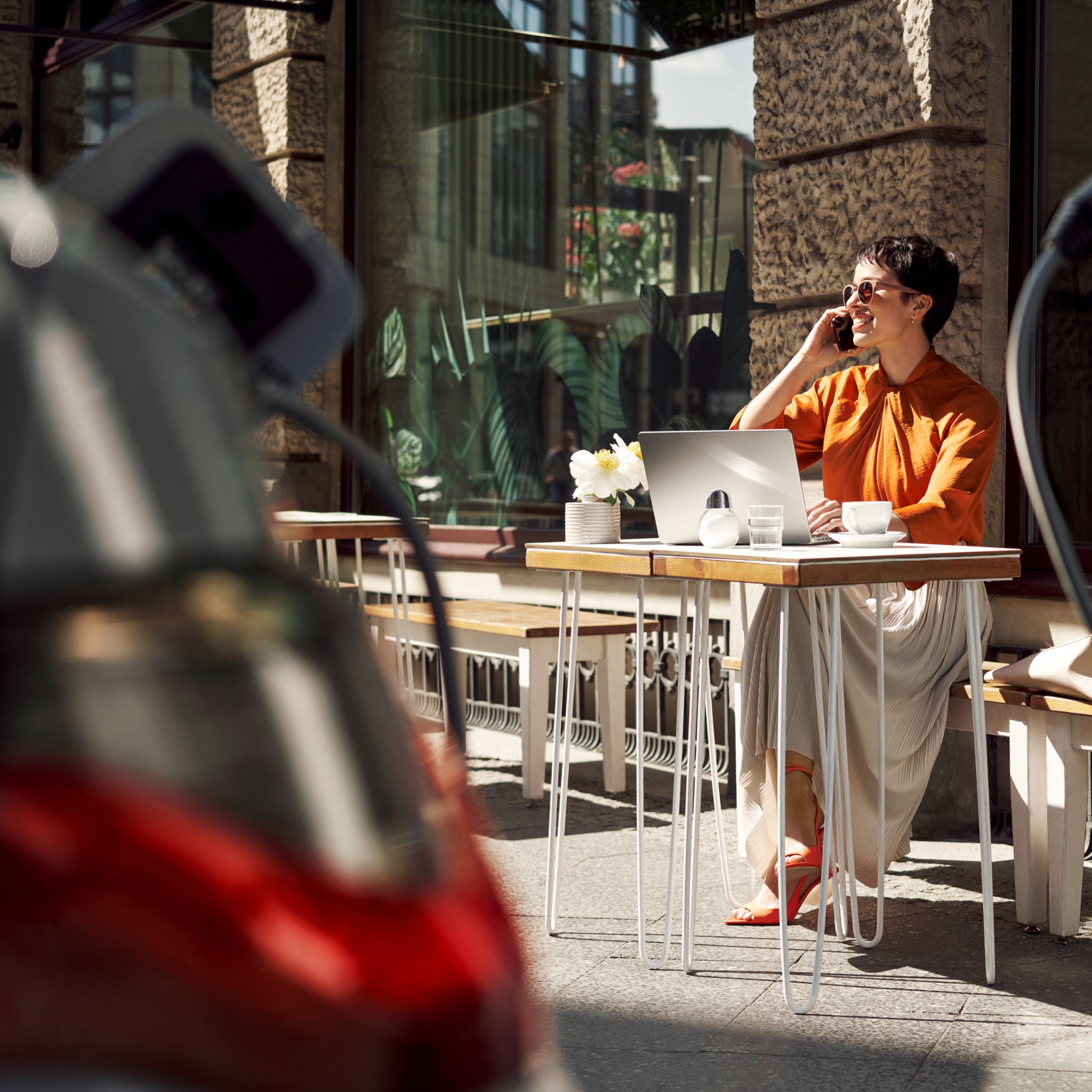 Žena sedí pri stole pred kaviarňou s otvoreným notebookom a telefonuje. V popredí sa nabíja BMW