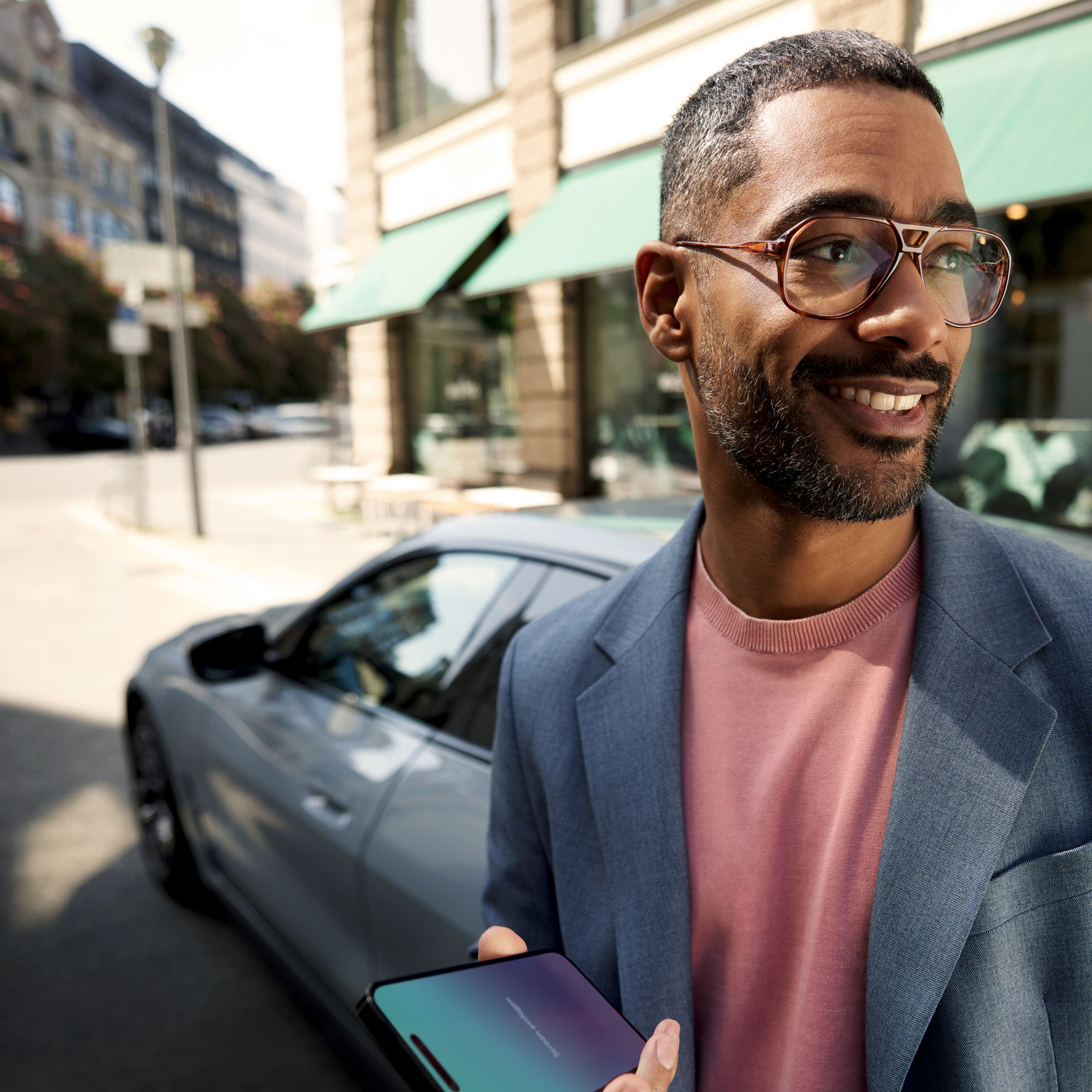 Muž držiaci smartfón stojí pred BMW, usmieva sa a pozerá sa doľava