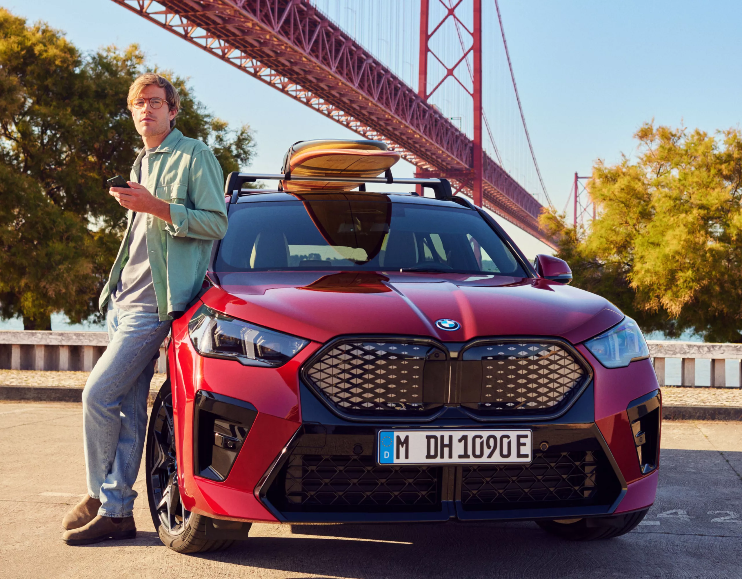 Ein Mann lehnt an seinem BMW, auf dessen Dach sich ein Surfbrett befindet. Dahinter ist eine Brücke über einem Fluss zu sehen
