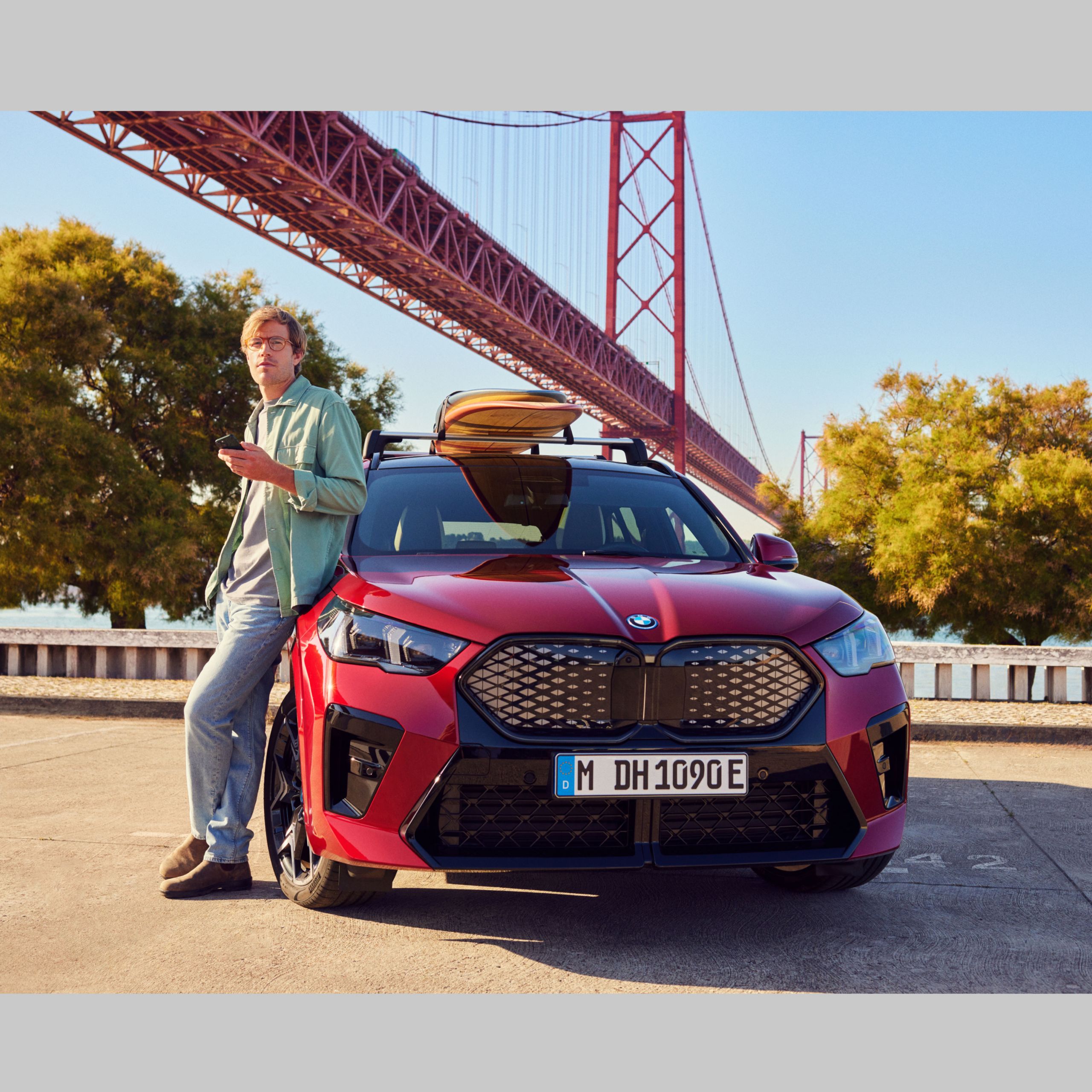 Muž sa opiera o svoje BMW, ktoré má na streche surf. V pozadí vidieť most cez rieku