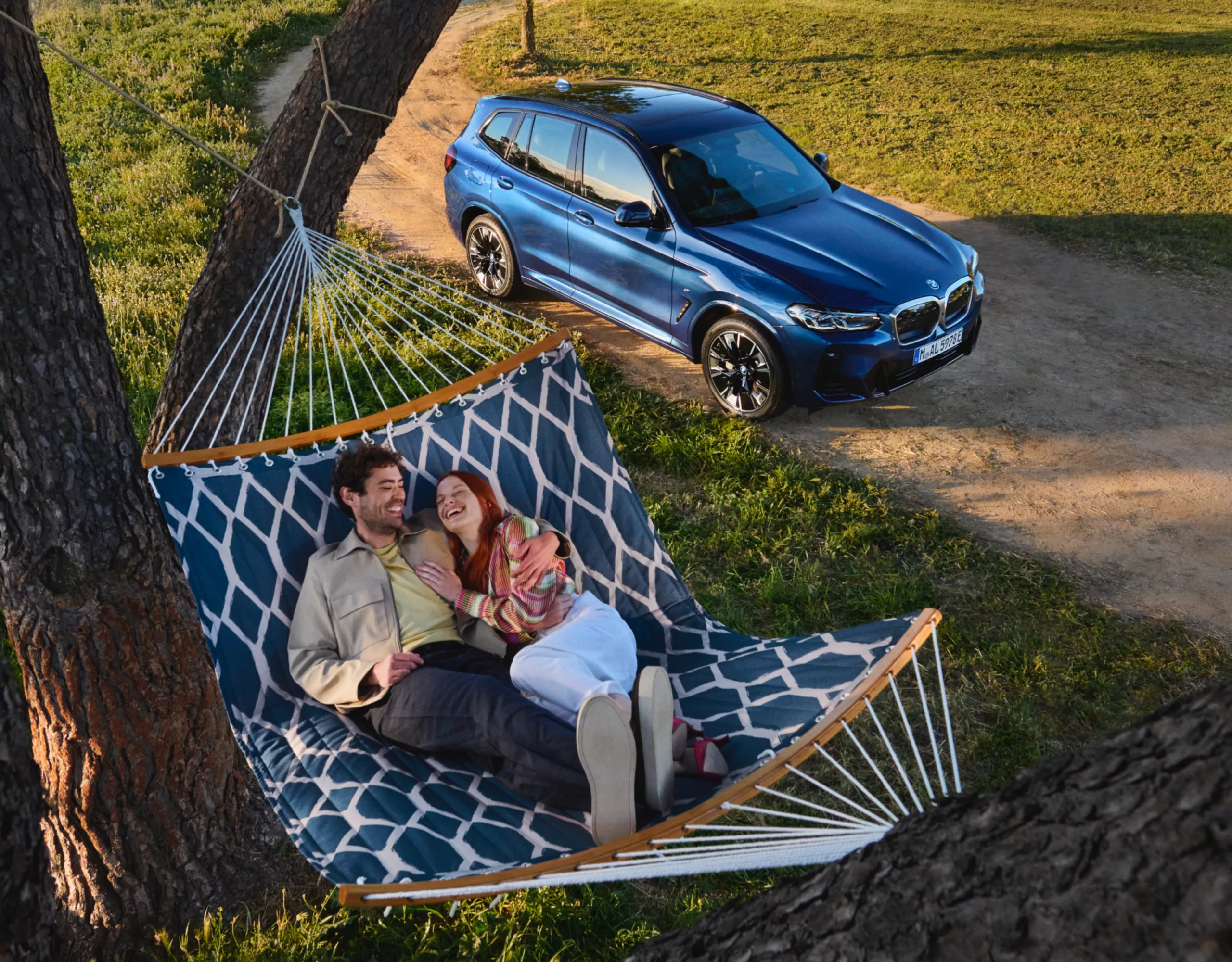 Ein Mann und eine Frau liegen kuschelnd in einer Hängematte, die zwischen zwei Bäumen in der Natur hängt. Neben ihnen steht ein BMW