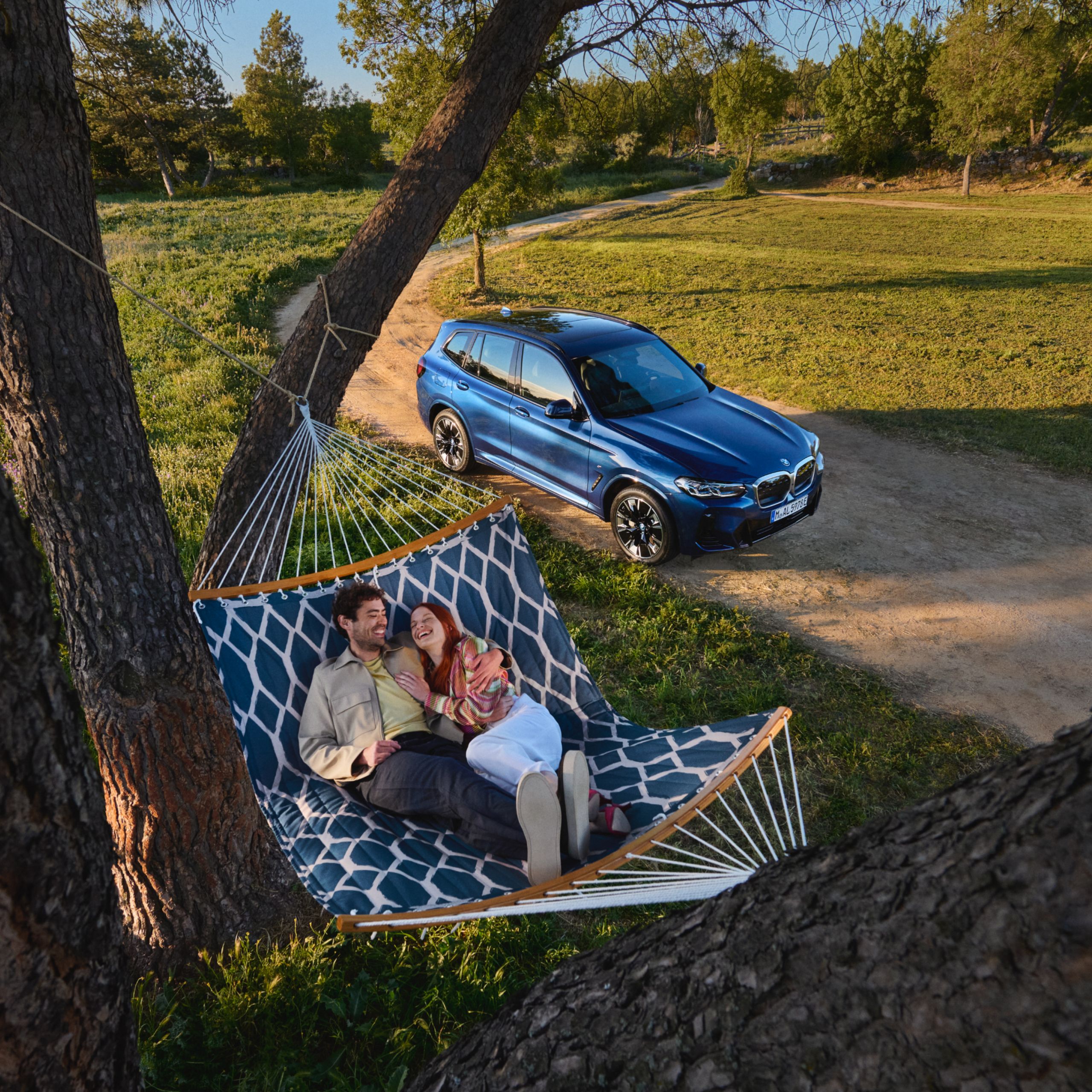 Muž a žena sa túlia vo visutom lôžku zavesenom medzi dvoma stromami v prírode. Vedľa nich stojí BMW