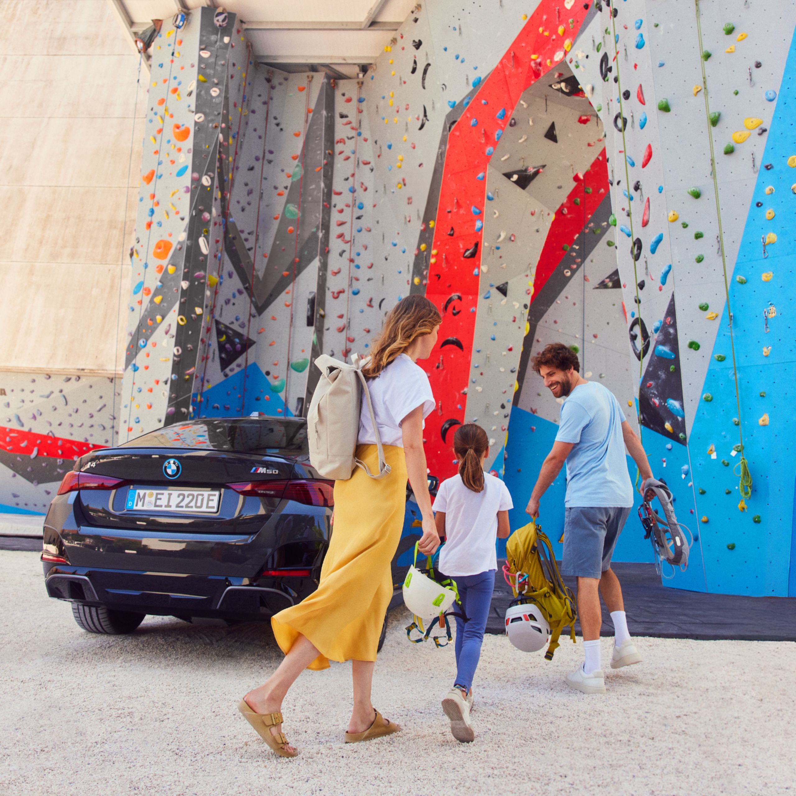 Skupina ľudí s batohmi a prilbami sa približuje k lezeckej stene. Vedľa nich je zaparkované BMW