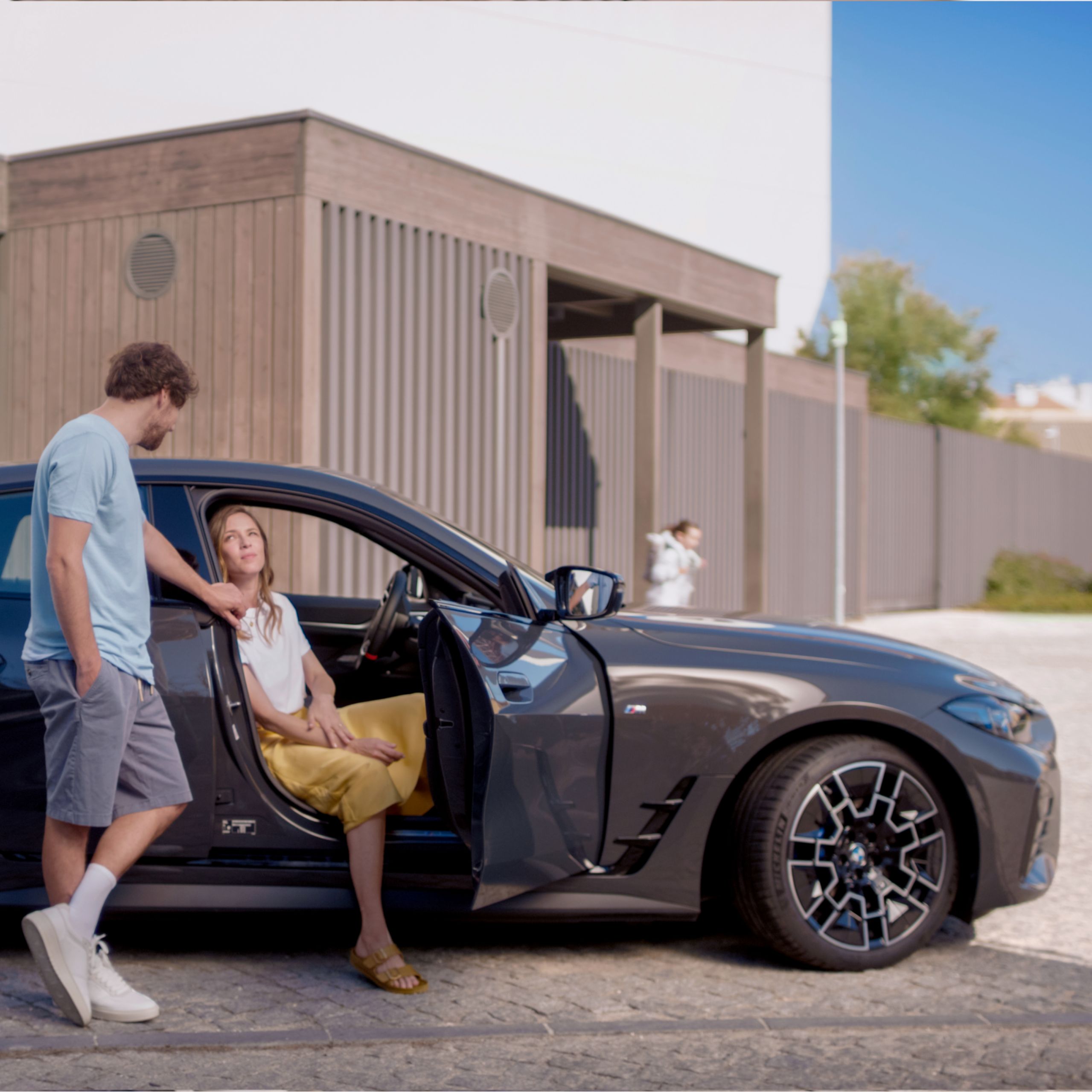 Žena sedí v BMW s otvorenými dverami. Muž sa opiera o vozidlo s domom viditeľným v pozadí