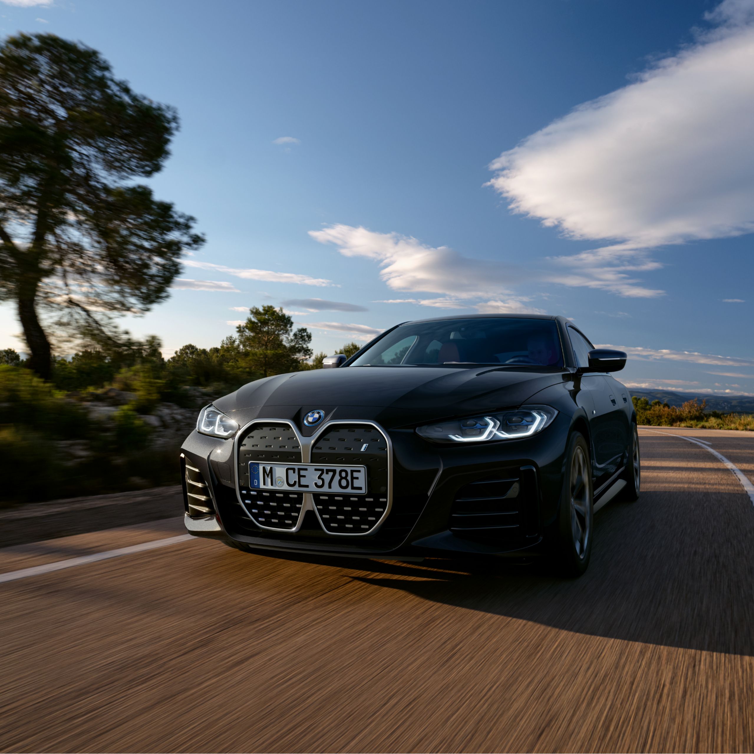 Μια BMW i4 κινείται σε δρόμο που περιβάλλεται από τη φύση και τον γαλάζιο ουρανό