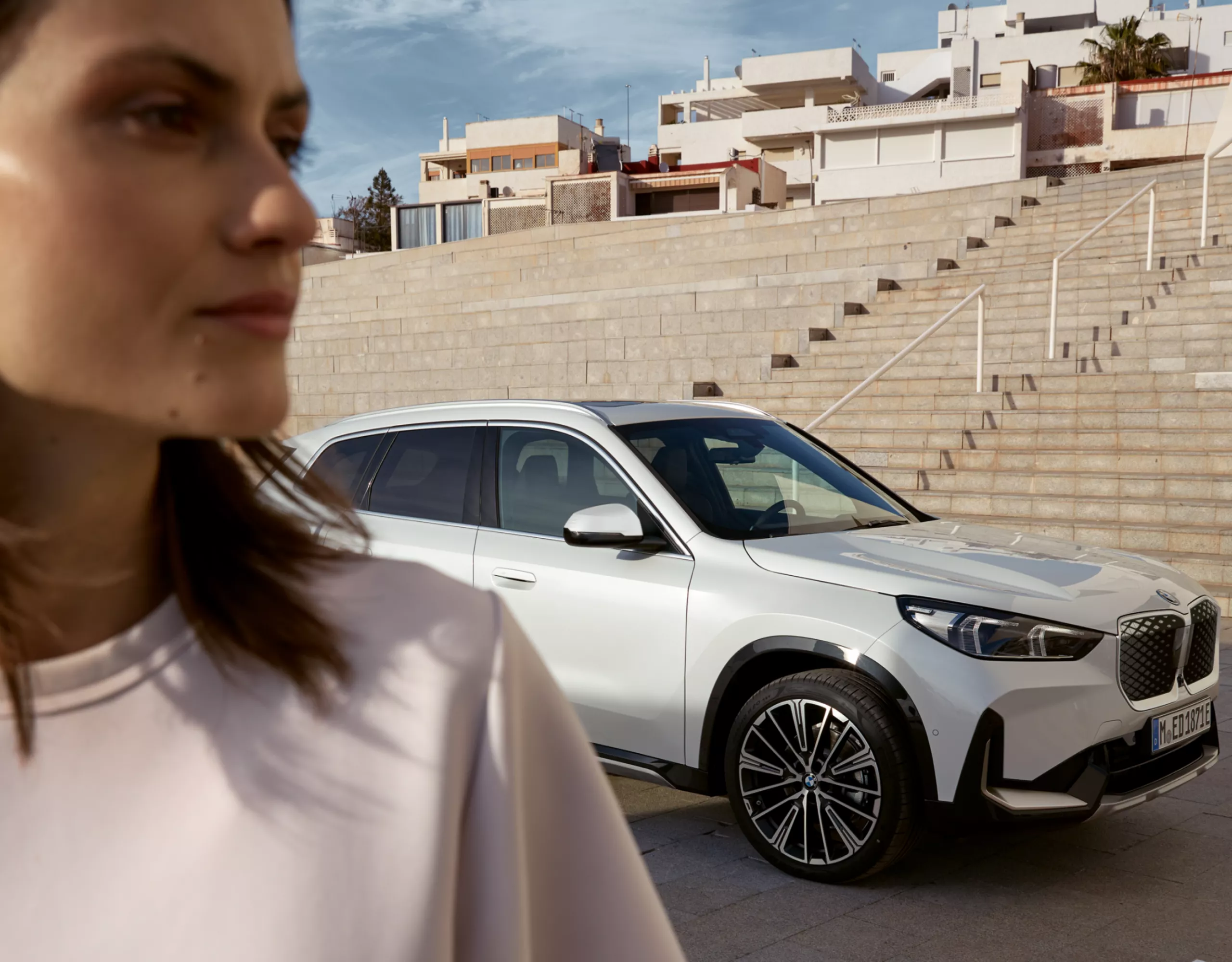 Μια γυναίκα στέκεται μπροστά από μια BMW. Πίσω της, πολλά σκαλοπάτια οδηγούν σε ένα κτίριο.