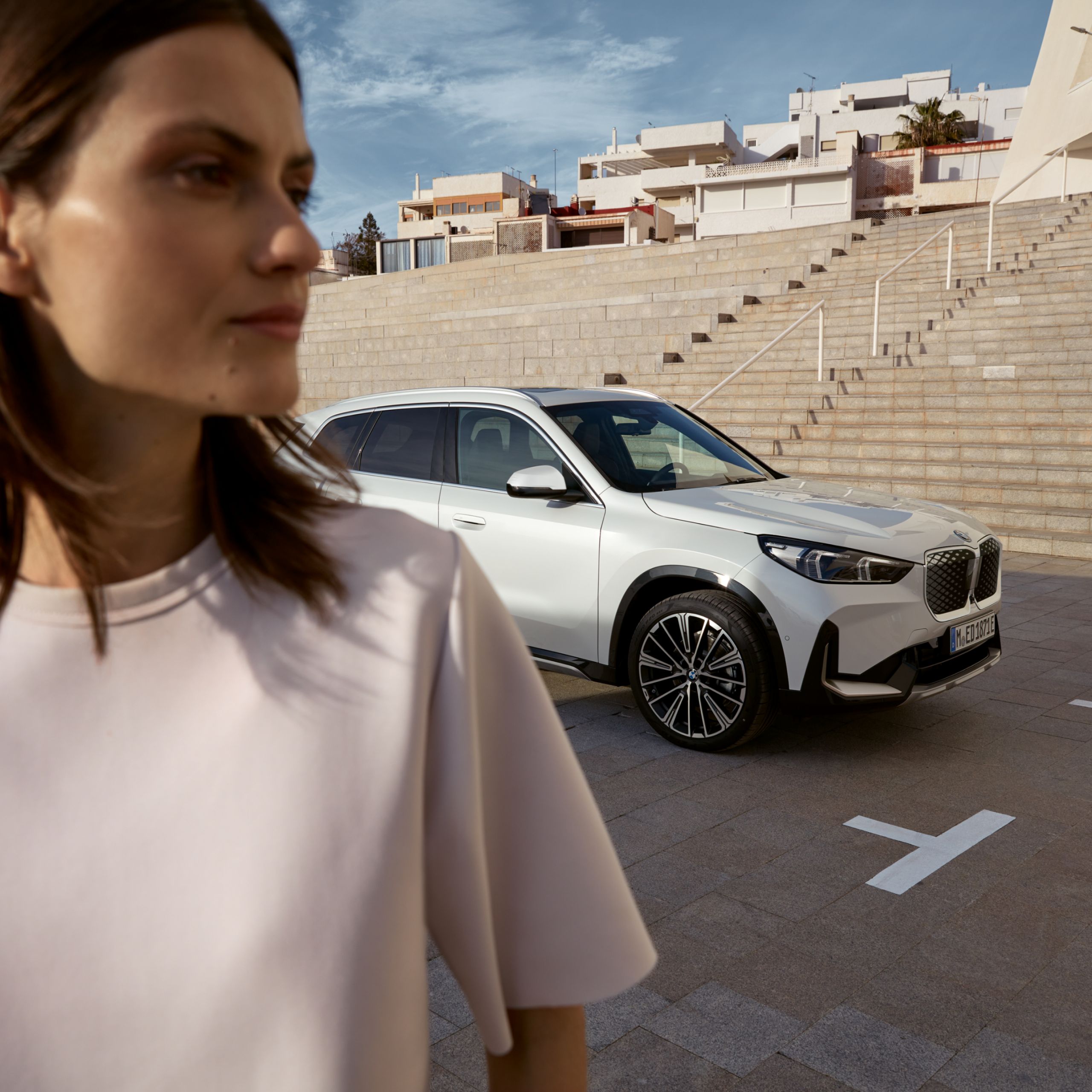 Μια γυναίκα στέκεται μπροστά από μια BMW. Πίσω της, πολλά σκαλοπάτια οδηγούν σε ένα κτίριο.