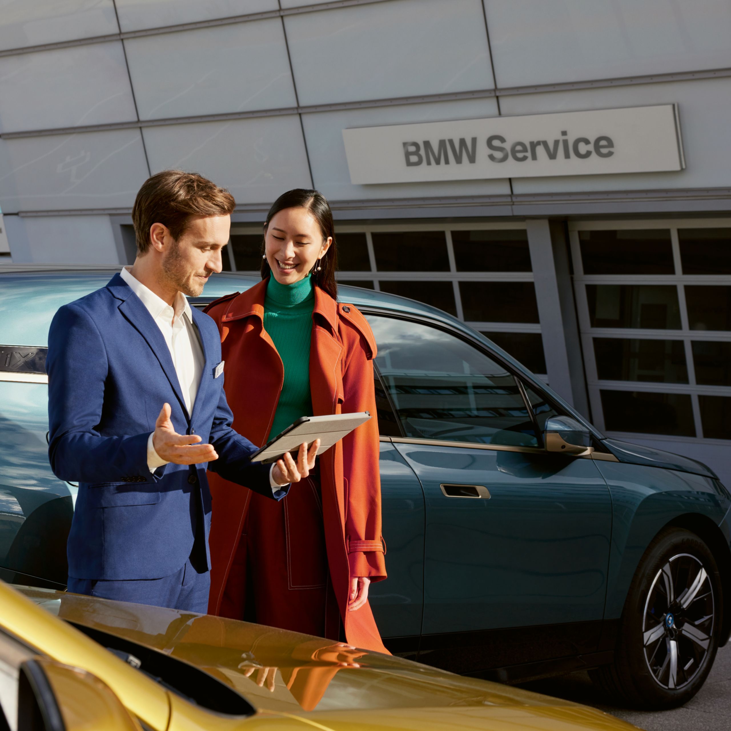 Ένας υπάλληλος στέκεται με ένα tablet δίπλα σε έναν πελάτη, και οι δύο κοιτούν τη συσκευή. Στο βάθος φαίνεται μια BMW και ένα κτίριο BMW σέρβις.