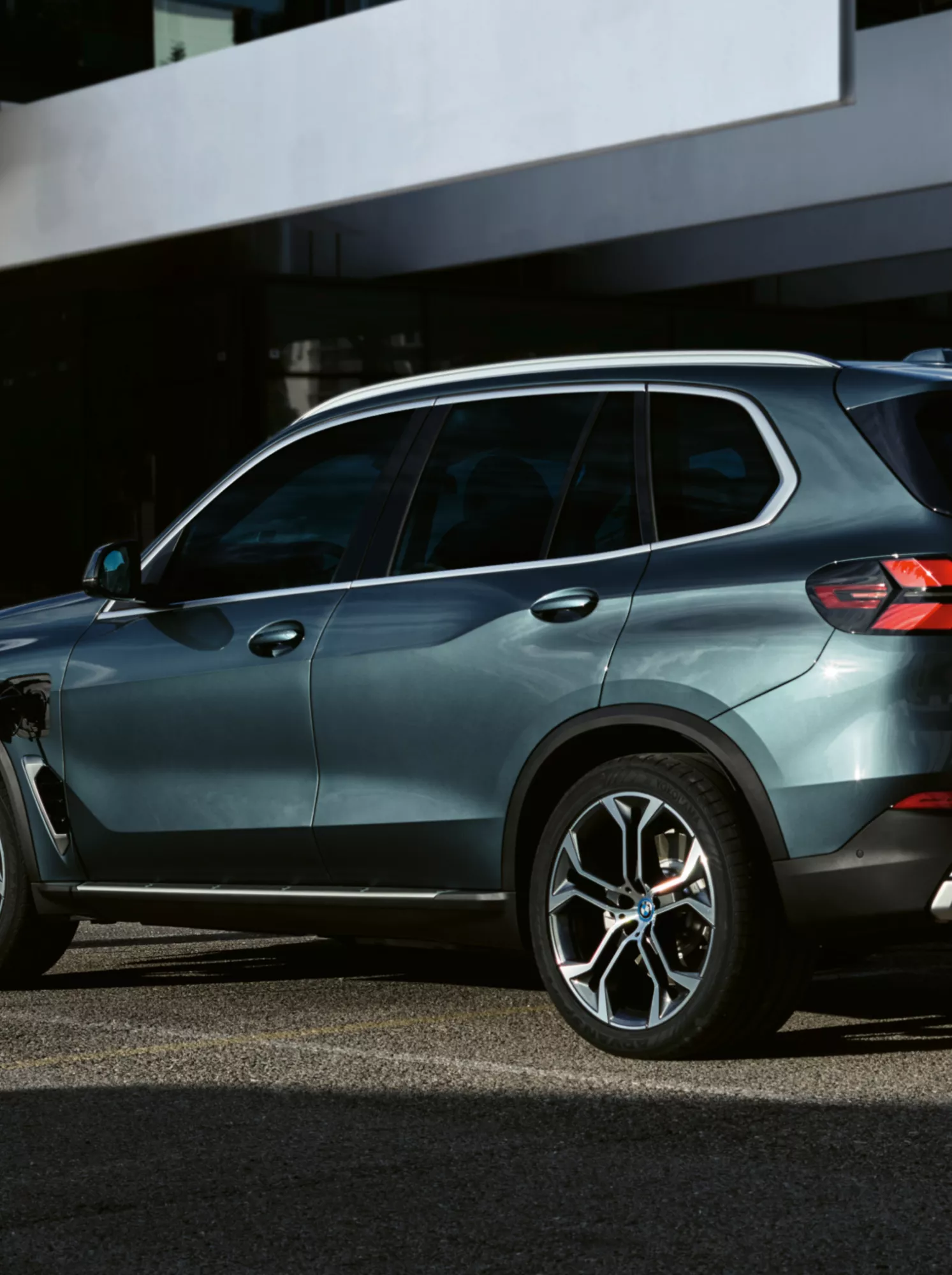 A steel blue BMW X5 plug-in hybrid SUV connected to a white EV charging station, shot from the rear three-quarter angle in front of a modern architectural building