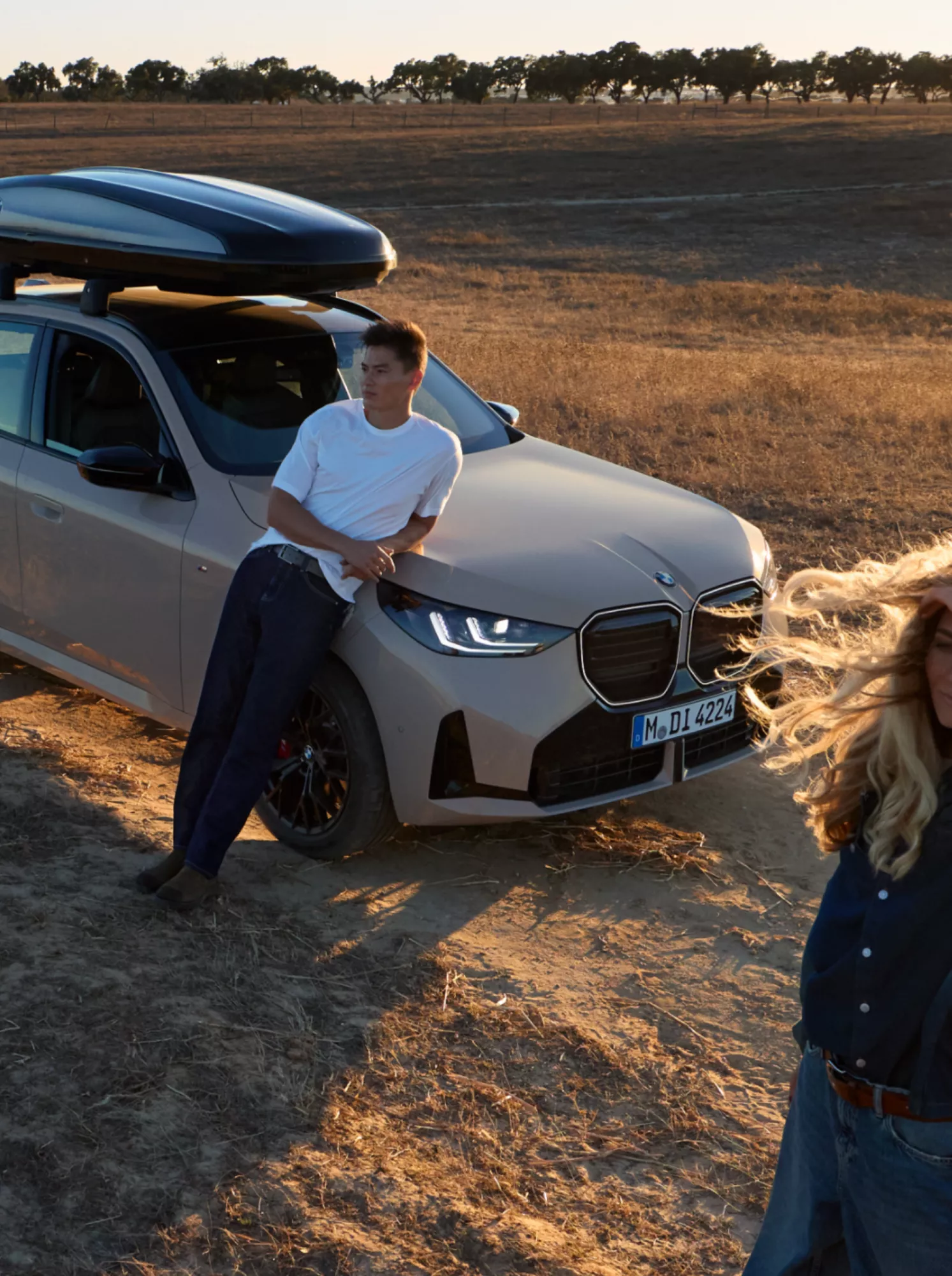 Ein BMW mit Dachbox steht im Sonnenschein auf freiem Feld, ein Mann lehnt sich an das Fahrzeug, eine Frau blickt in die Ferne