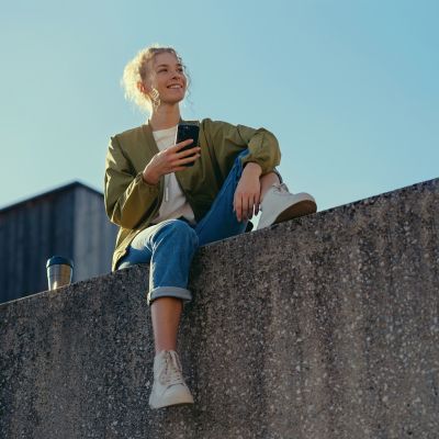 A woman smiles while sitting on a ledge on her phone