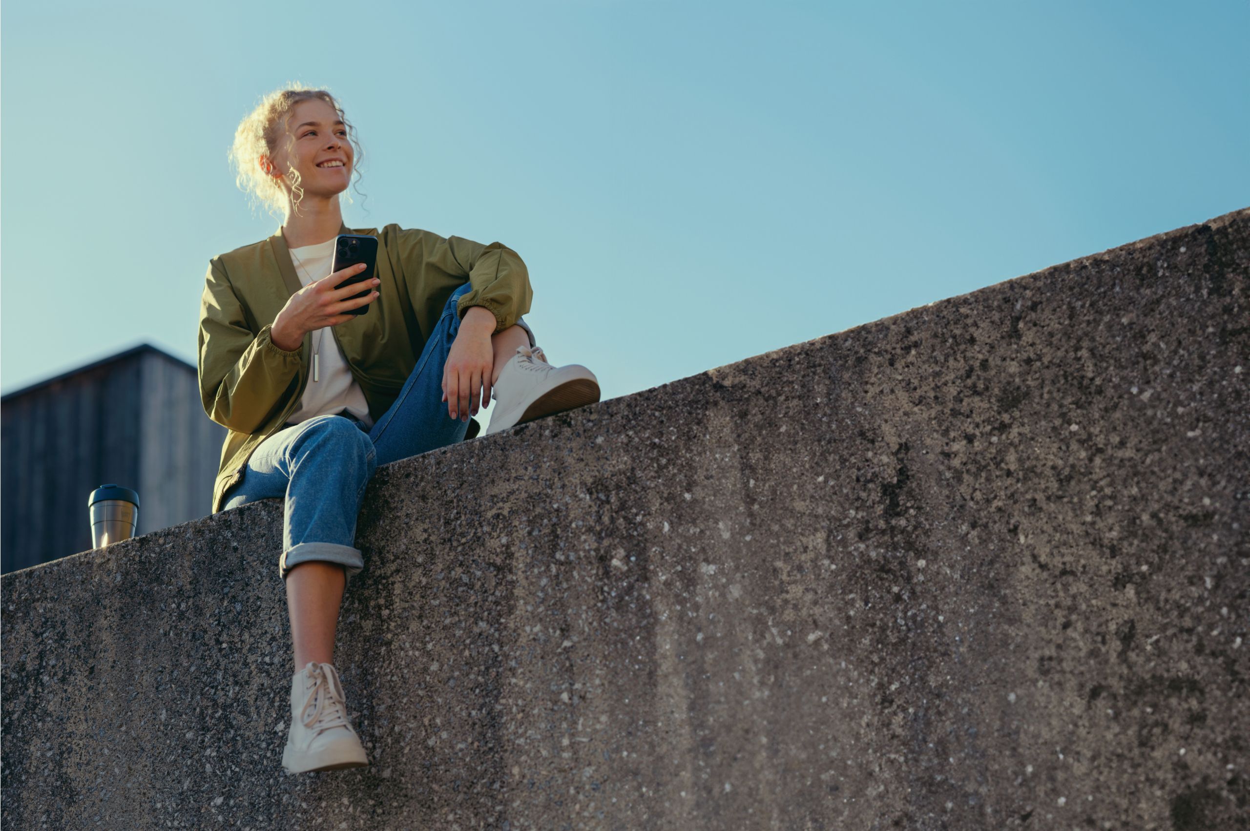 A woman smiles while sitting on a ledge on her phone