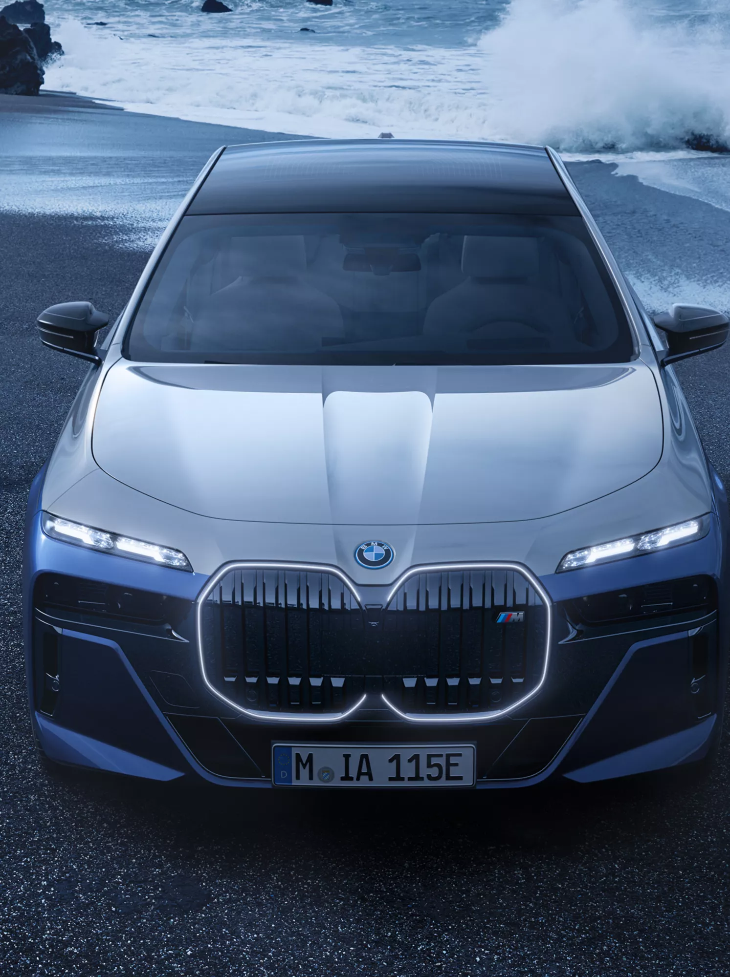 BMW i7 M70 xDrive auf einem Sandstrand vor einer felsigen Küste mit Brandung
