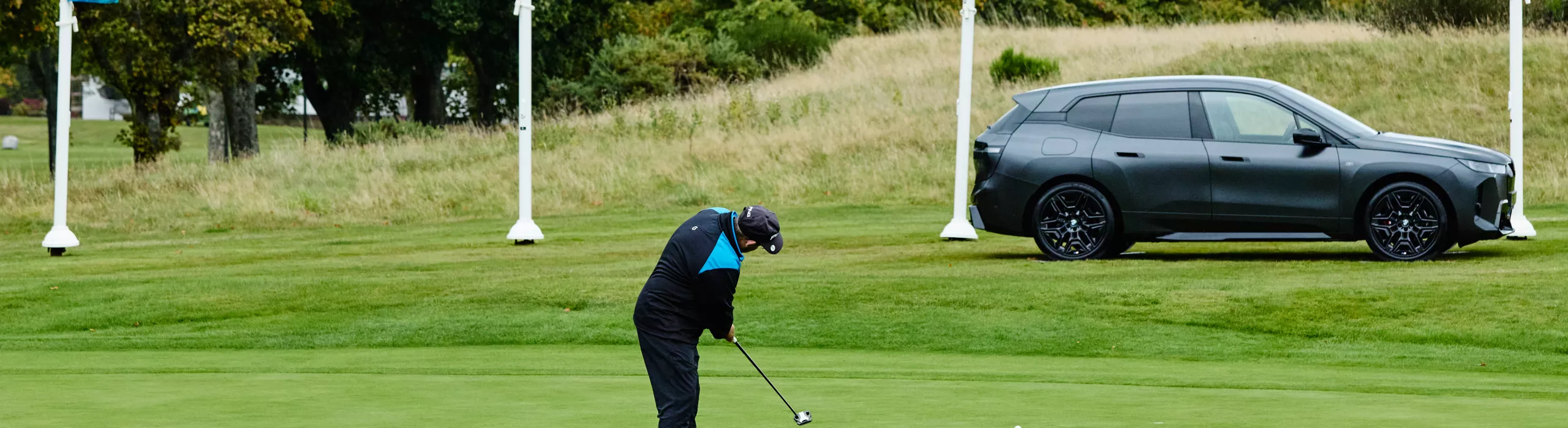Golfer mid-swing on an expansive green course during BMW Golf Cup National Final. Two others watch. Landscape of rolling hills and trees in background.