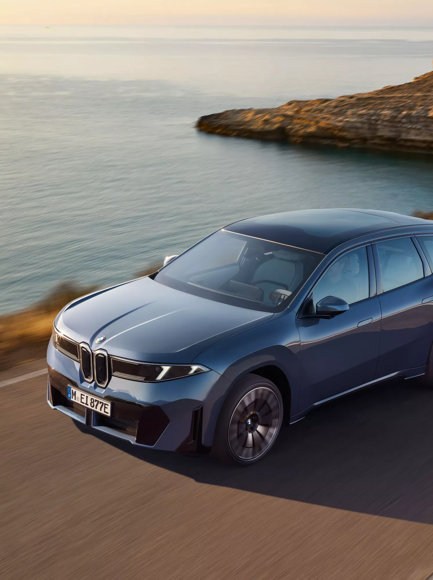 En blå BMW iX3 sett från sidan kör på en kustväg