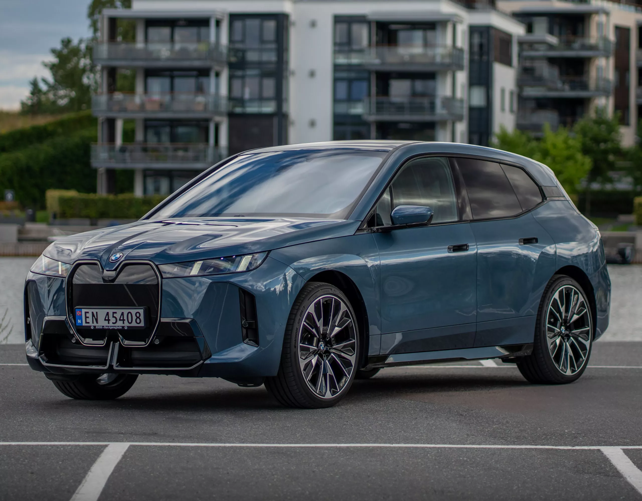 En BMW iX parkerad vid vatten med byggnader och grönska i bakgrunden.