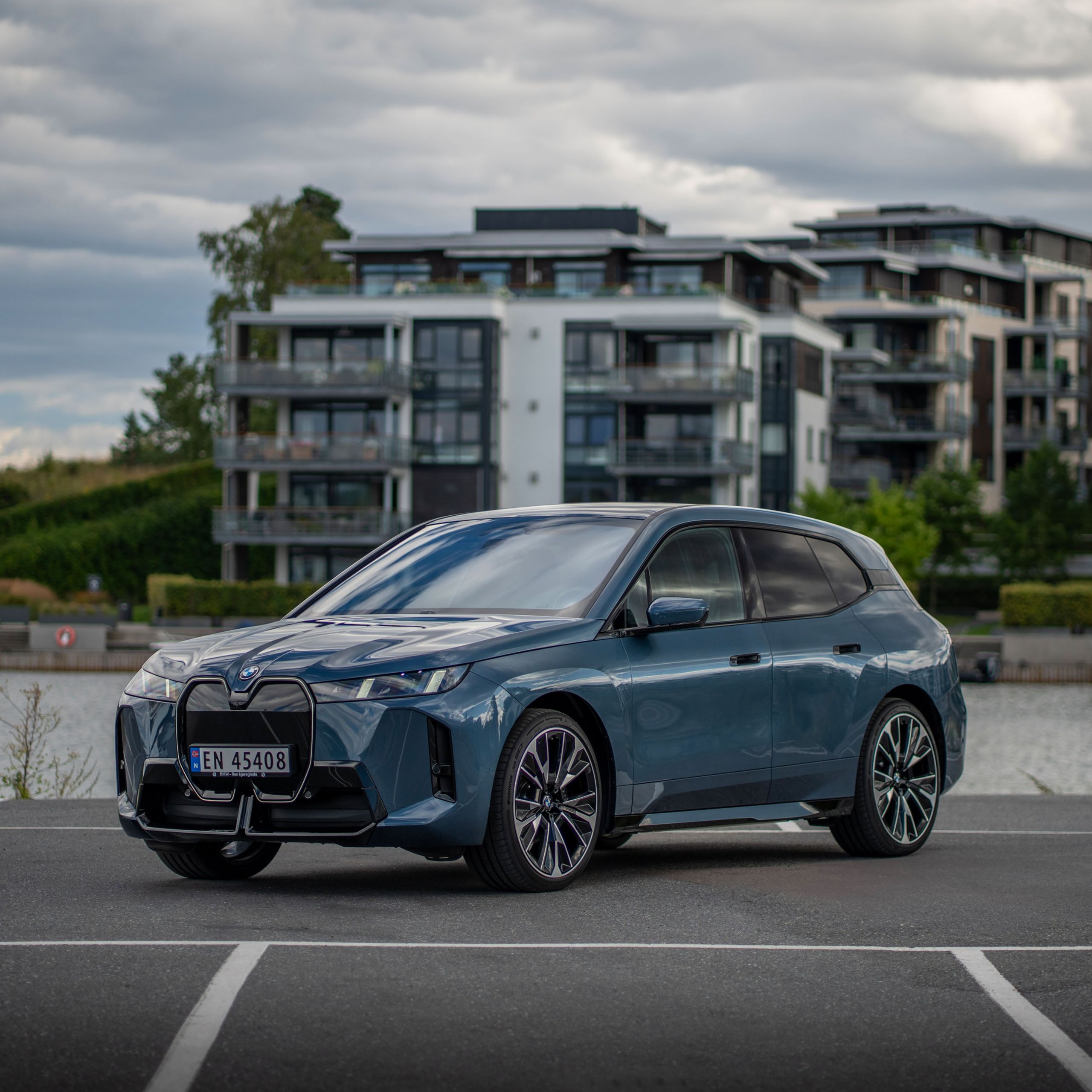En BMW iX parkerad vid vatten med byggnader och grönska i bakgrunden.