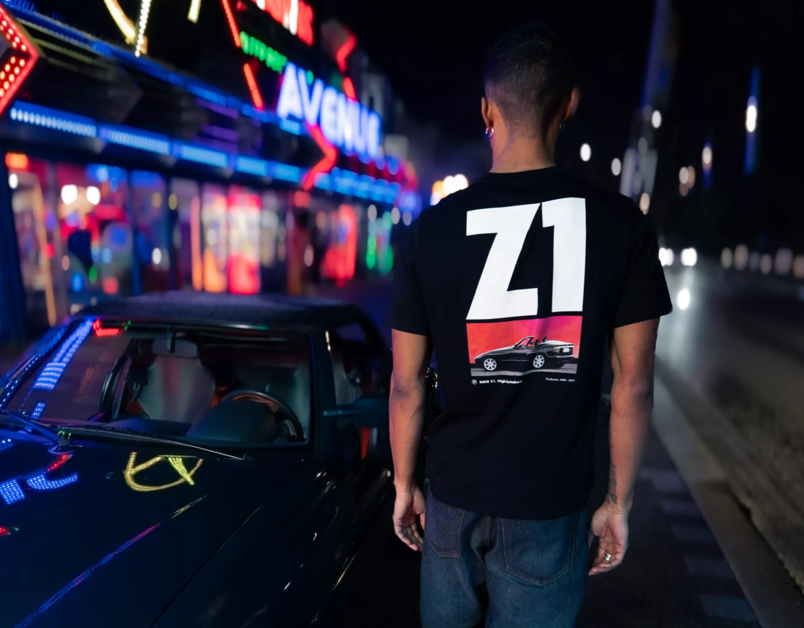 T-shirt BMW Lifestyle de couleur noire avec une grande inscription Z1 blanche dans le dos, et BMW Z1 de couleur noire sur fond rouge comme toile de fond