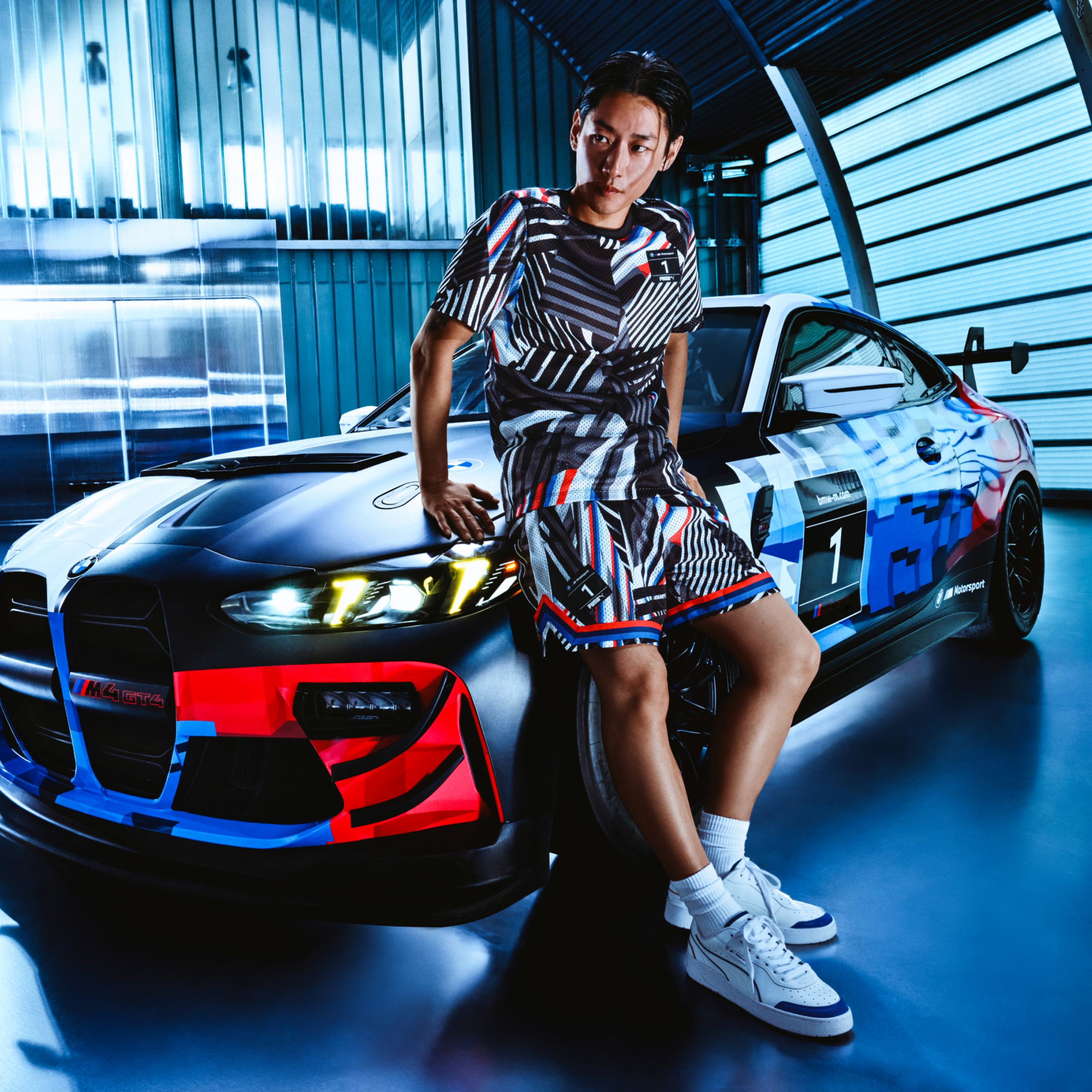BMW Lifestyle Oblečení BMW M Sport s trikem a šortkami v klasických barvách BMW M, které nosí mladý muž opírající se o BMW M4