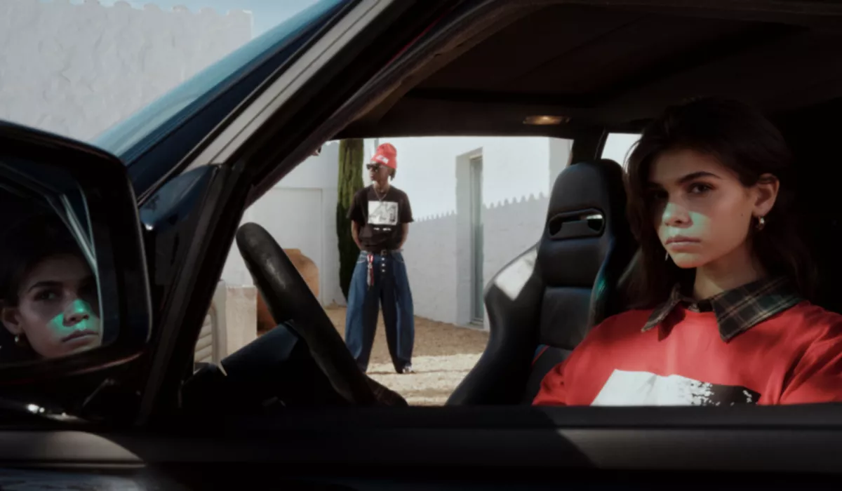 หญิงสาวสวมเสื้อคอลเลคชัน BMW M นั่งอยู่หลังพวงมาลัย BMW หญิงสาวยืนอยู่ด้านหลัง มองเห็นได้ผ่านประตูฝั่งผู้โดยสาร โดยสวมเสื้อและหมวกบีนนี่จากคอลเลคชัน BMW