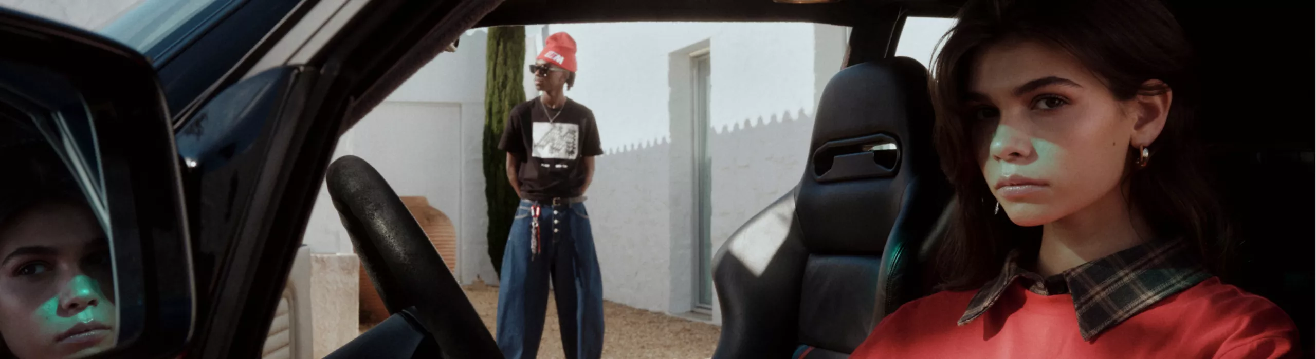 A young woman in a BMW M collection shirt sits at the wheel of a BMW. A young woman stands in the background visible through the passenger door, also wearing a BMW collection shirt and beanie