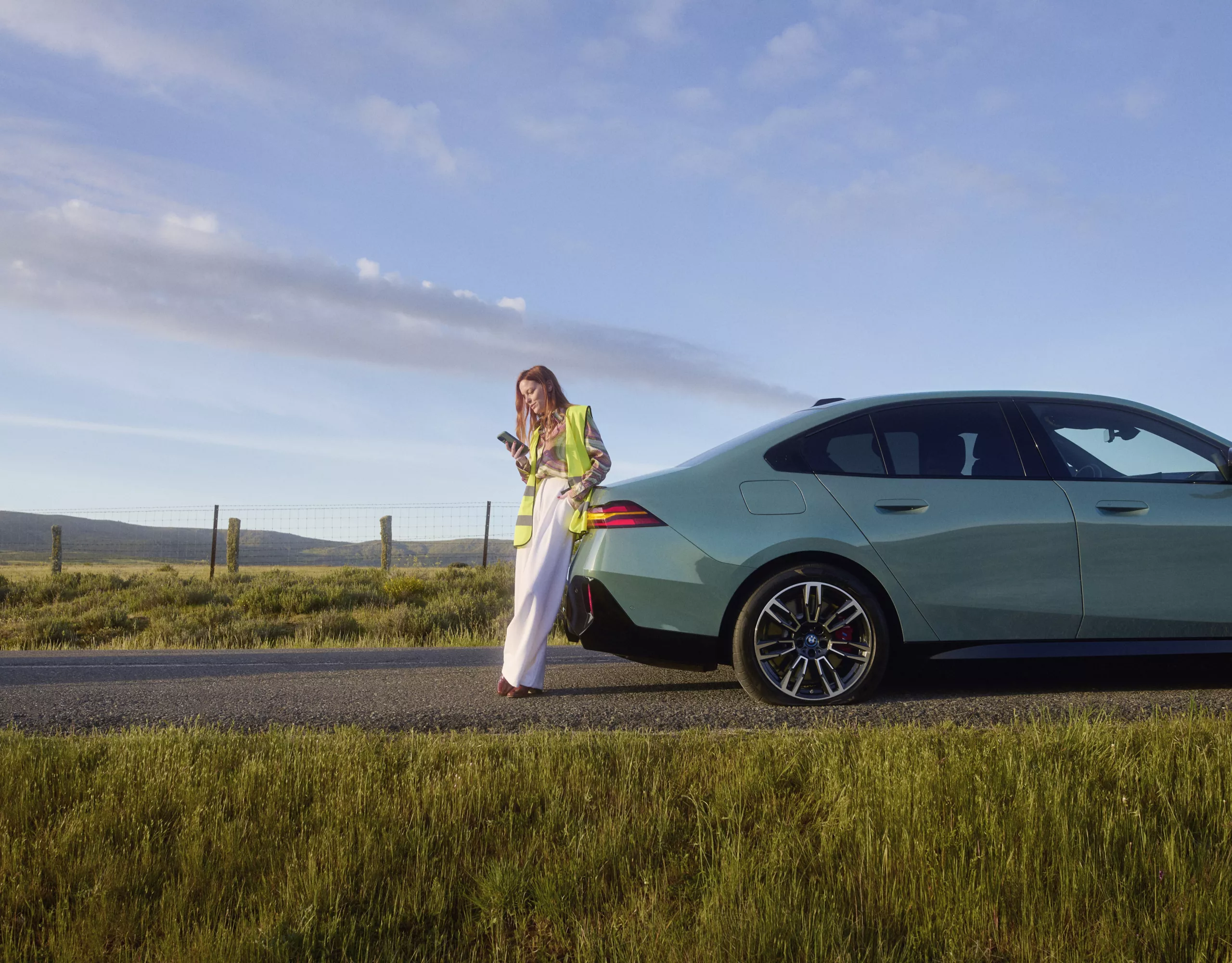 A woman sits in a BMW with the door open. A man leans on the vehicle with a house visible in the background