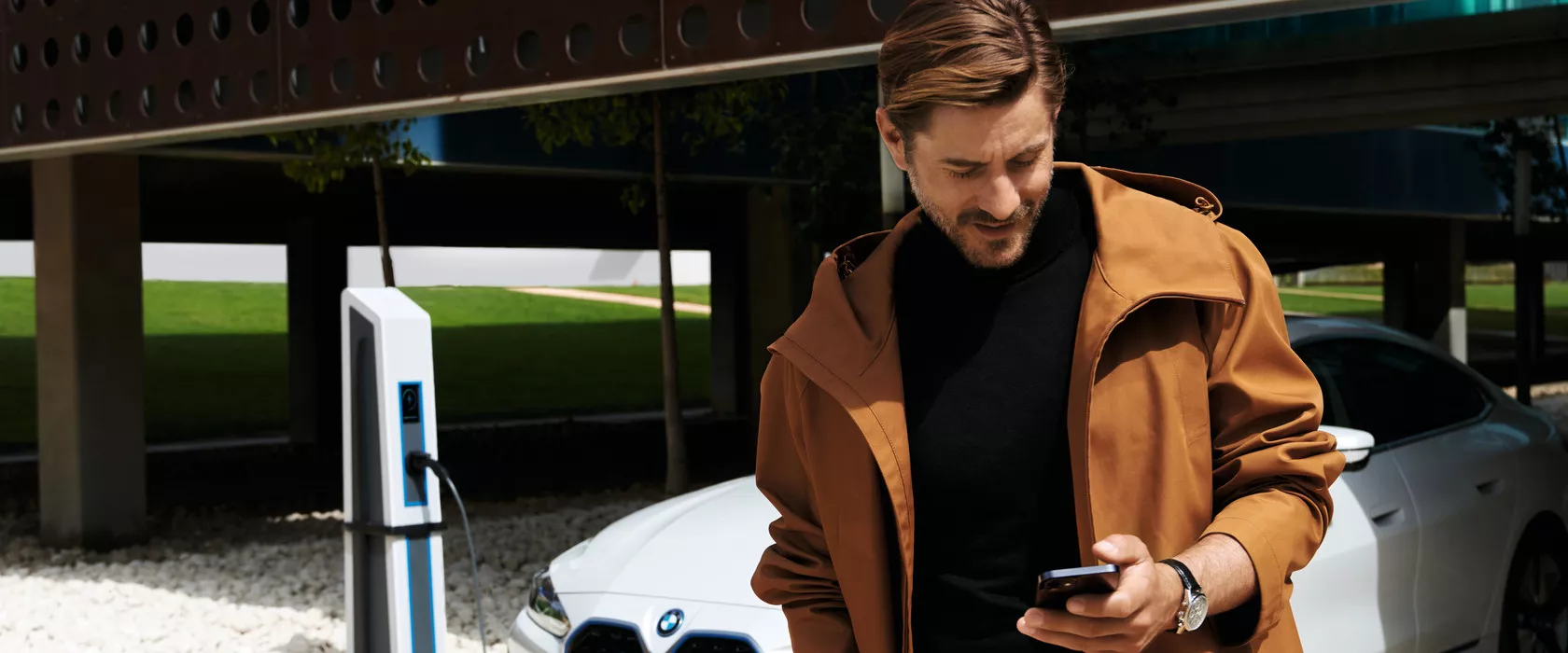 Hombre usando su teléfono móvil junto a un BMW conectado a un cargador eléctrico.
